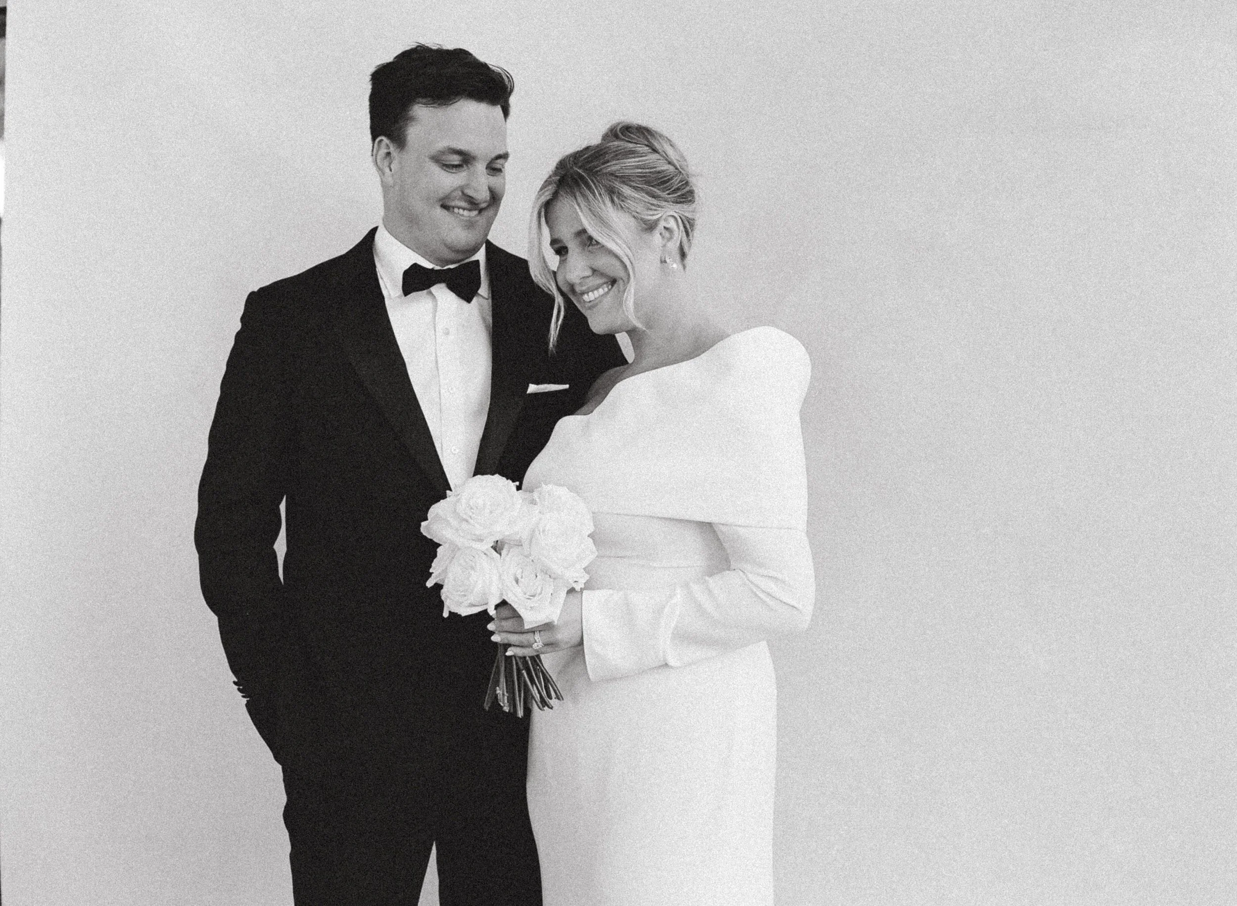 A black-and-white photo of a bride and groom on their wedding day. The groom is wearing a tuxedo and the bride is in a white wedding dress holding a bouquet of flowers. They are smiling and looking at each other, standing against a plain background.