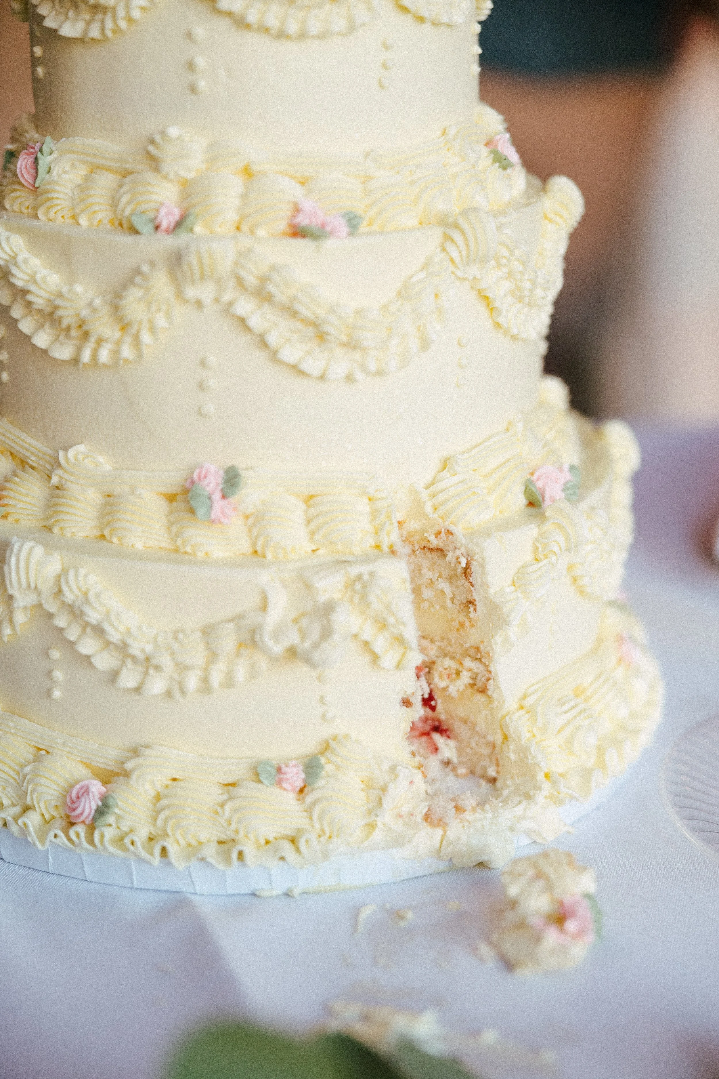 Close-up of a multi-tiered white wedding cake decorated with piped cream and small pastel-colored sugar flowers, with a piece cut out showing the inside.