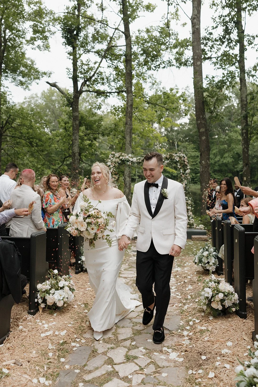 A bride and groom walking hand in hand down an outdoor wedding aisle through a wooded area, smiling happily, with guests celebrating and throwing flower petals around them.