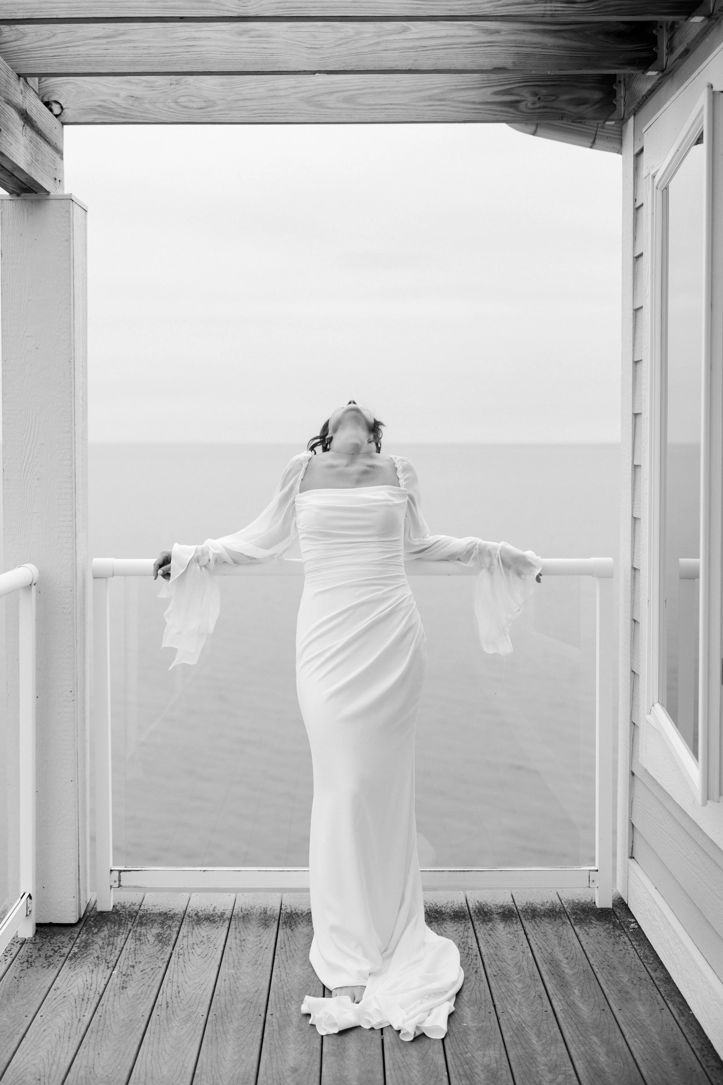 A woman in a white dress stands on a wooden deck, with her arms resting on a railing, her head tilted back, and her face turned upward. The scene is framed by an open doorway, with the sky and horizon visible in the background.