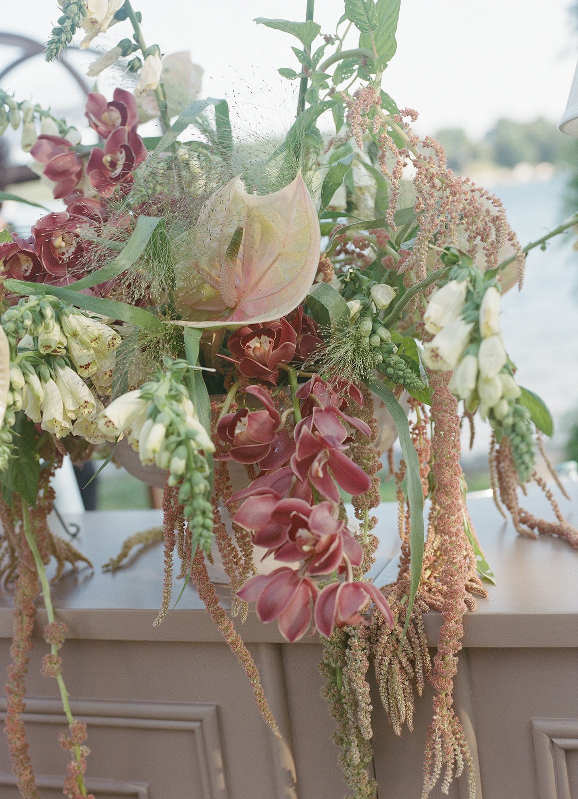 A floral arrangement with pink orchids, white flowers, and greenery on a table, with a body of water and trees in the background.