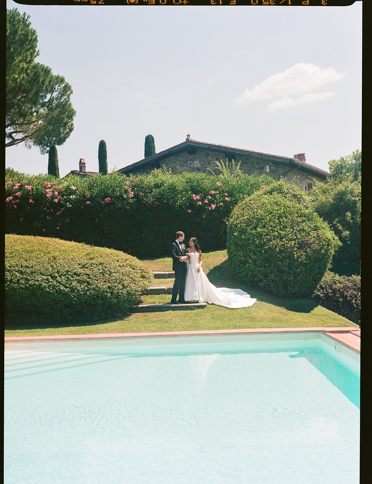 A bride and groom in wedding attire standing by a pool in a garden with green bushes and trees on a sunny day.