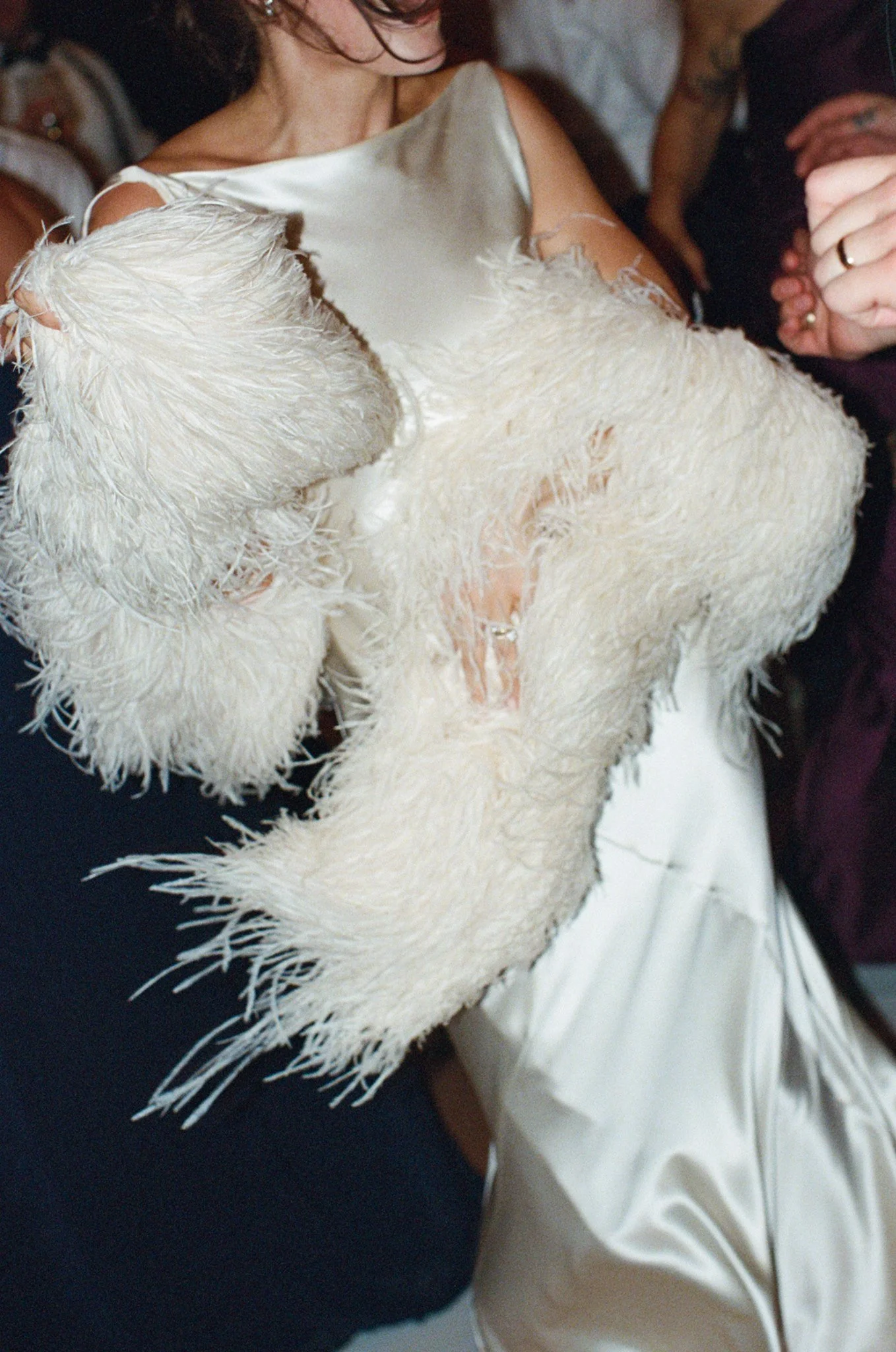 Close-up of a person wearing a white, textured dress with feathers on the sleeves, surrounded by other people at an event.