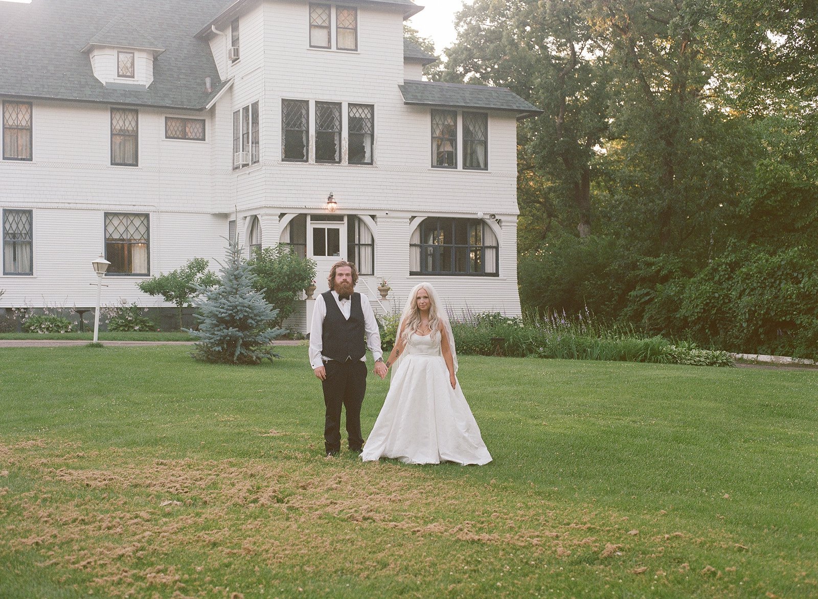 A bride and groom standing on a lawn in front of a large white house, holding hands. The groom has a beard and wears a black vest over a white shirt. The bride has long blonde hair and wears a white wedding dress.