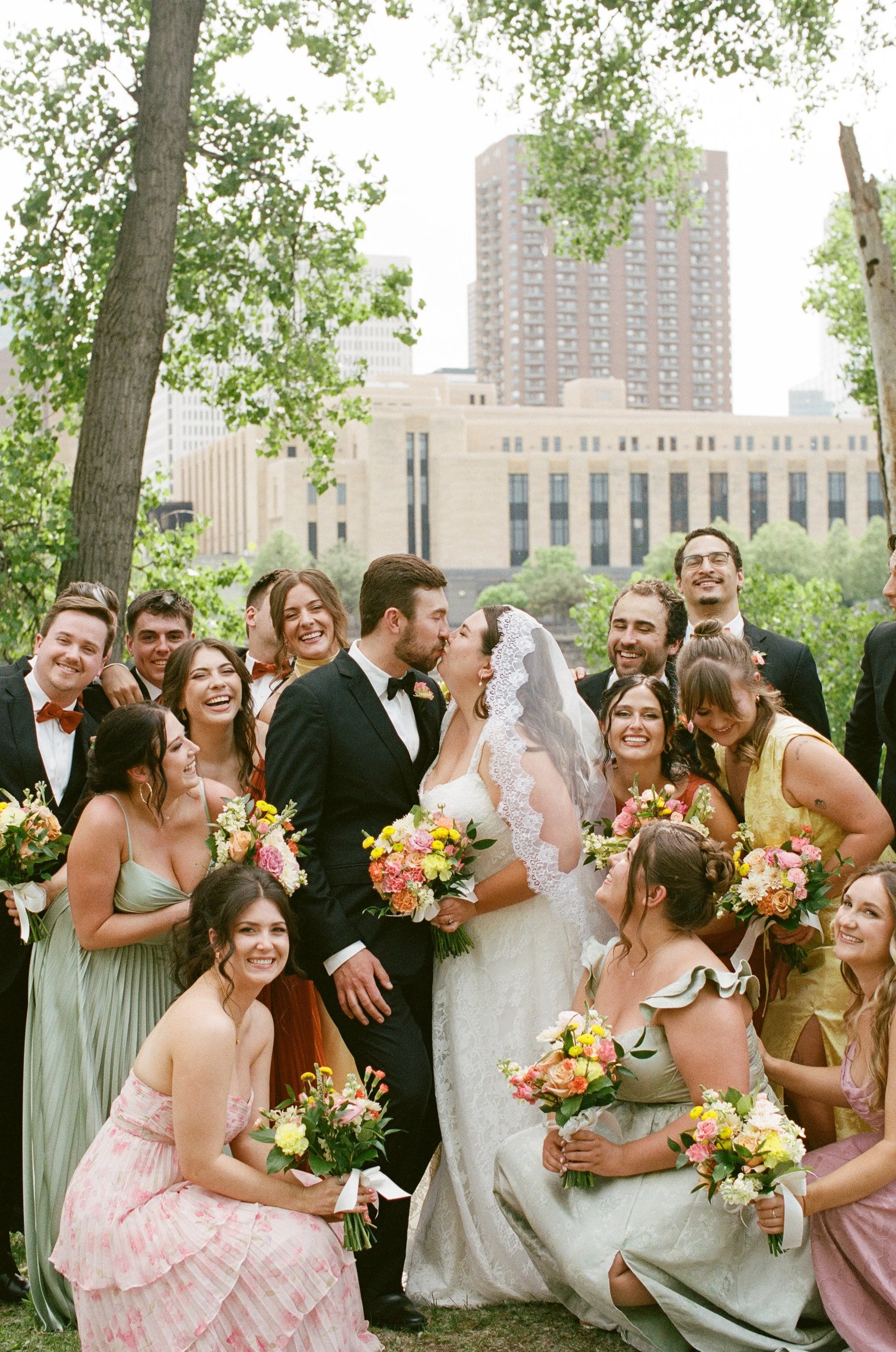 A group of people at a wedding, with some holding bouquets of flowers, standing outdoors in front of trees and tall buildings, celebrating and smiling.