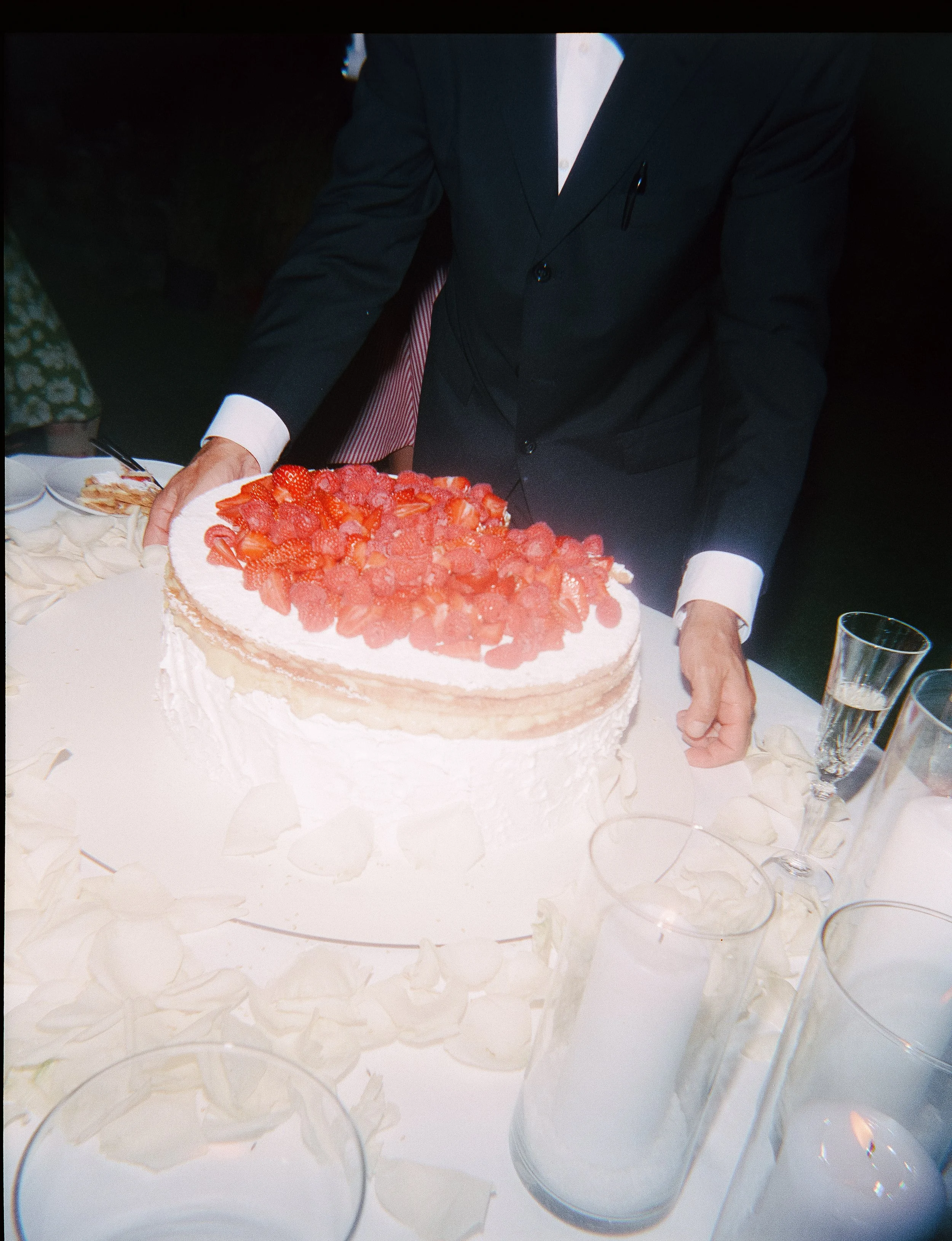 Man in a dark suit cutting a large strawberry shortcake cake at a celebration event.