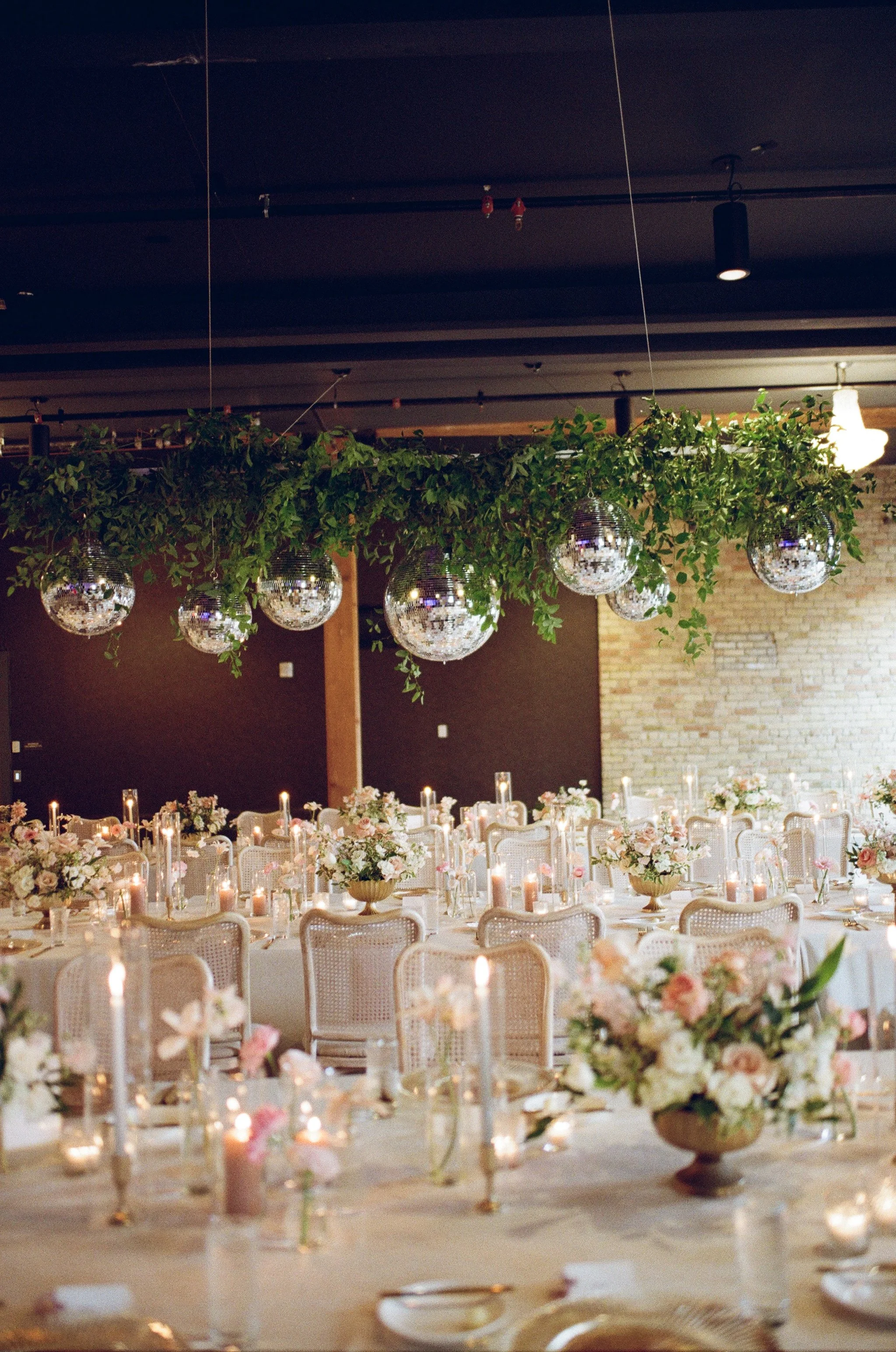 Elegant indoor wedding reception setup with long banquet tables, floral arrangements, candles, and hanging disco ball decorations, with warm ambient lighting and a brick wall in the background.