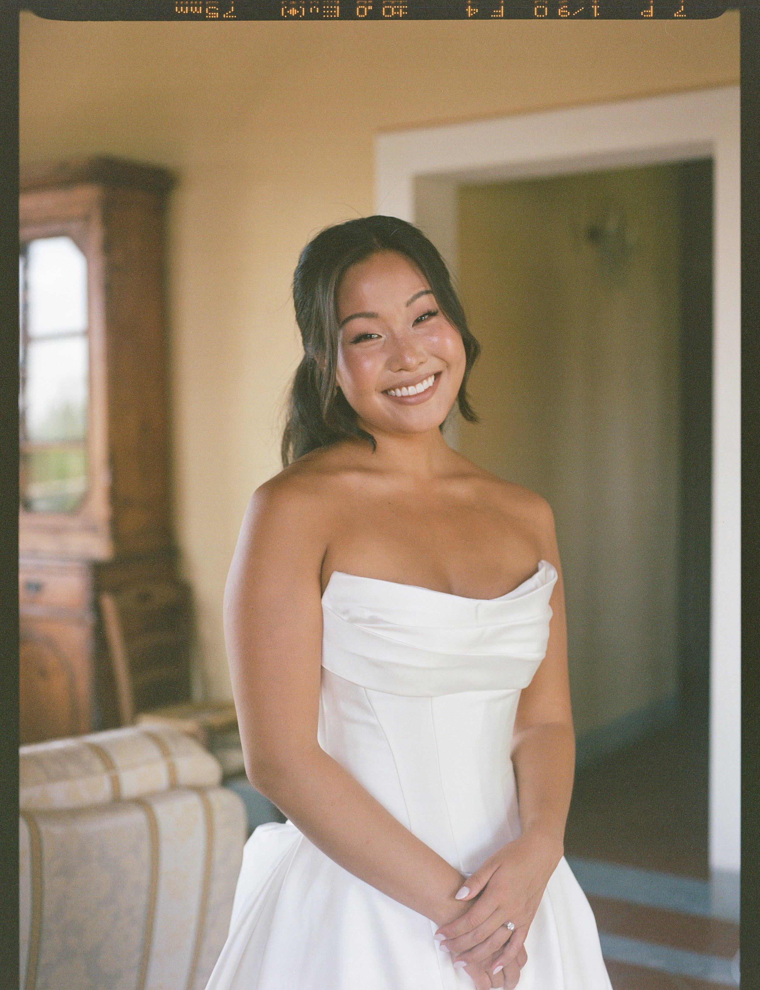 A woman smiling in a white strapless dress, standing indoors near a bed, with a wooden dresser and a window in the background.