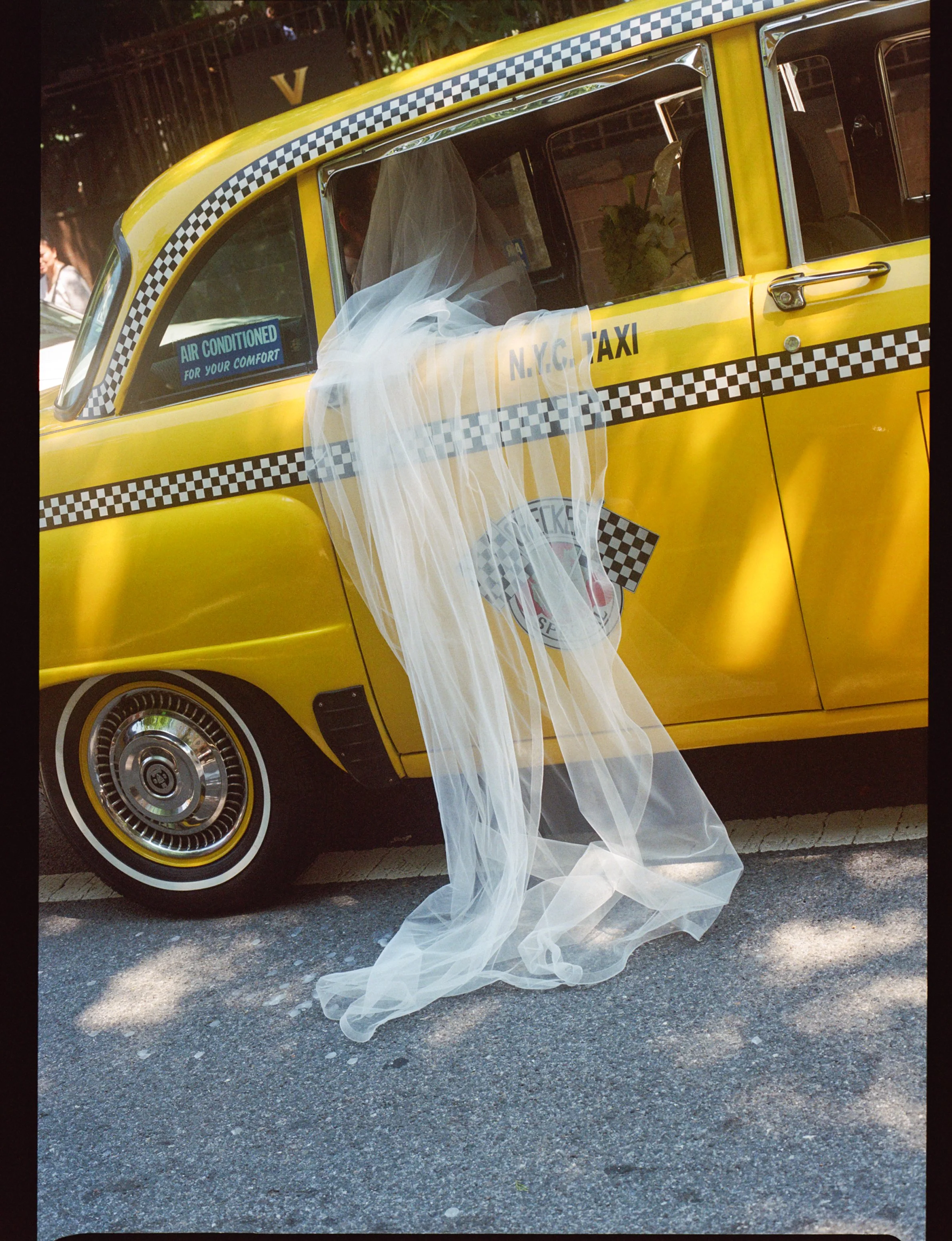 A yellow New York City taxi cab decorated with a white wedding veil hanging out of the window, with a sticker promoting air conditioning for comfort, parked on the street.