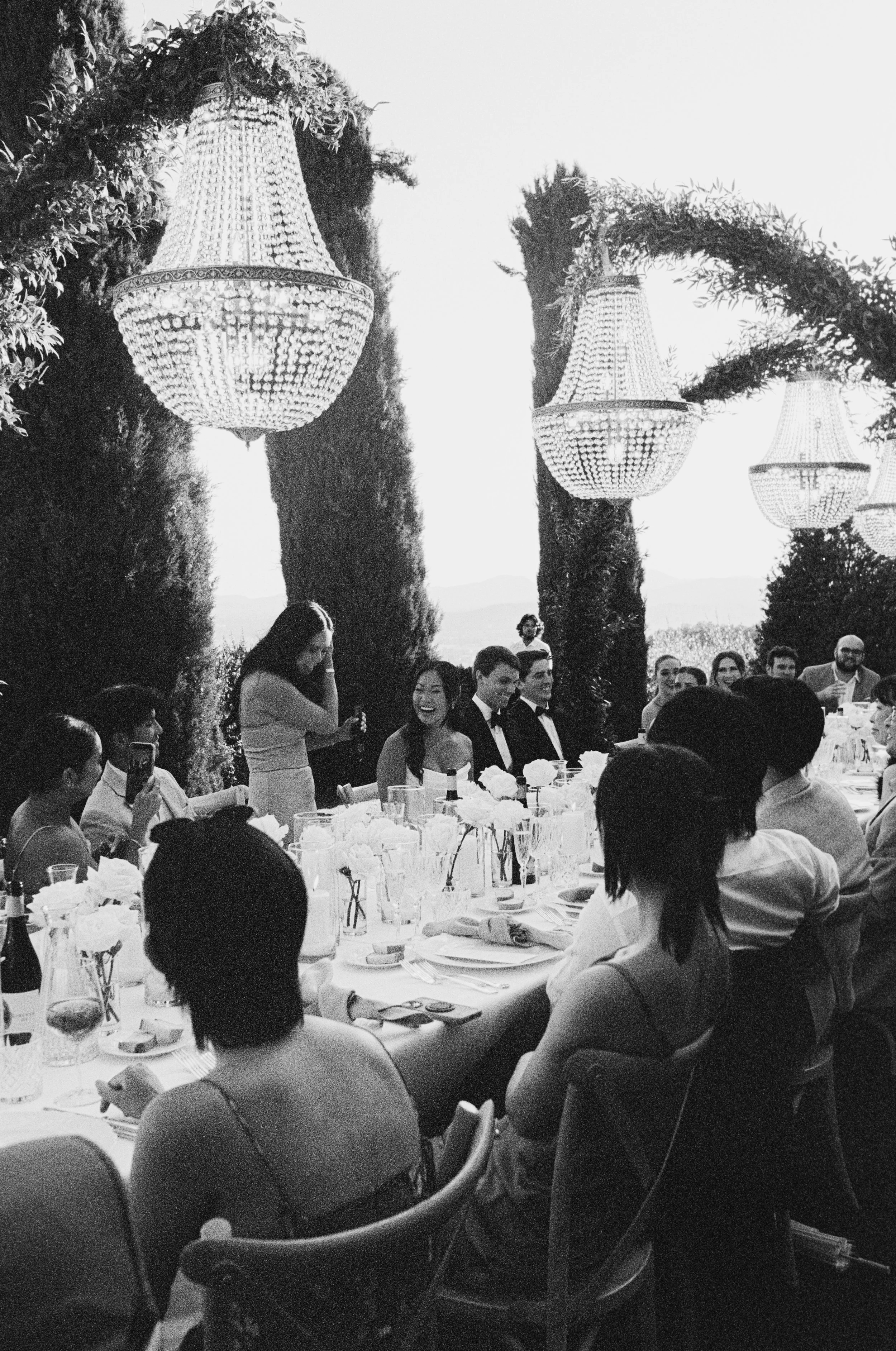 People sitting at a banquet table outdoors, smiling and talking. There are large chandeliers hanging from trees in the background.