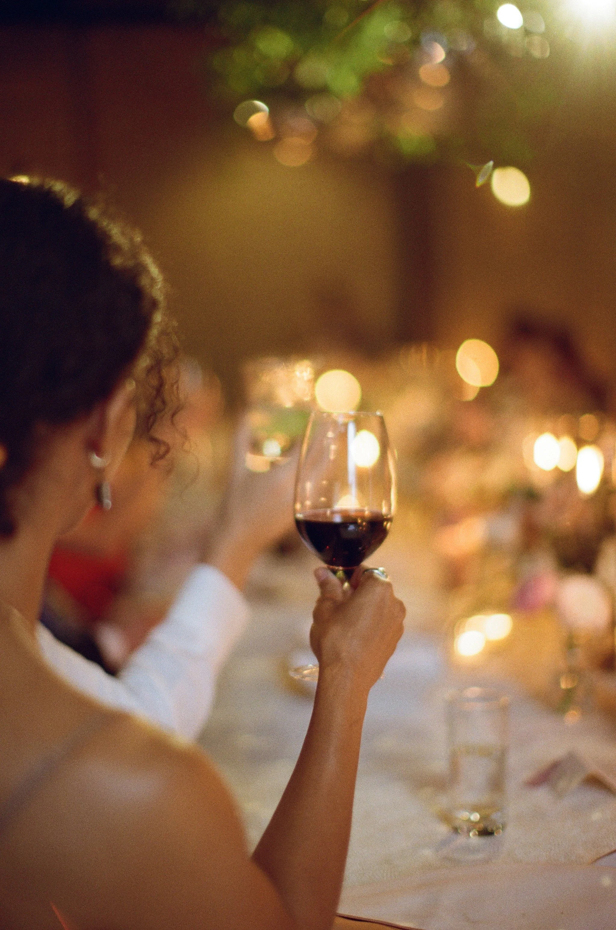 A woman with curly hair and earrings holding a glass of red wine at a social event with blurred warm lighting and other guests in the background.