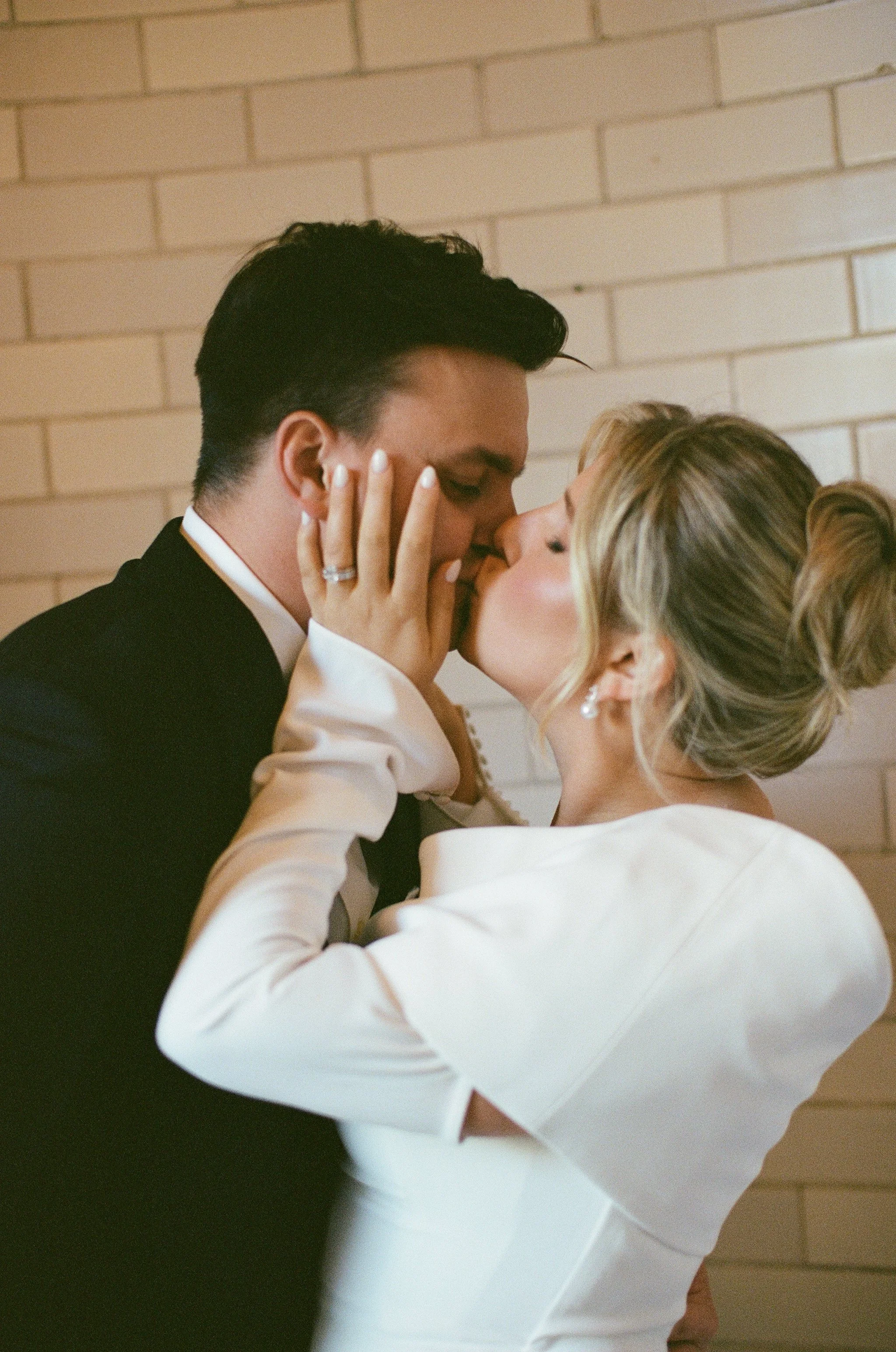 A couple, dressed in wedding attire, sharing a kiss with their eyes closed, against a brick wall background.