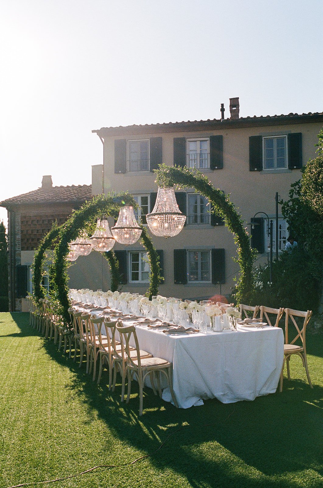 Long banquet table with white tablecloth, set with glasses, plates, and silverware, under decorative chandeliers and green arches, on a lawn in front of a beige house with black shutters.