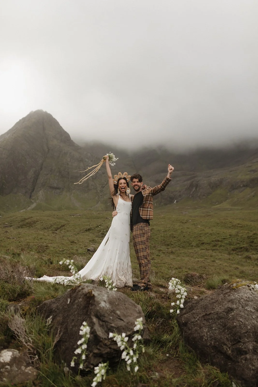 A newlywed couple celebrating outdoors in a foggy, mountainous landscape. The woman wears a white wedding dress and veil, holding a bouquet, smiling with her arm raised. The man wears a plaid suit, smiling, with one arm around the woman and the other