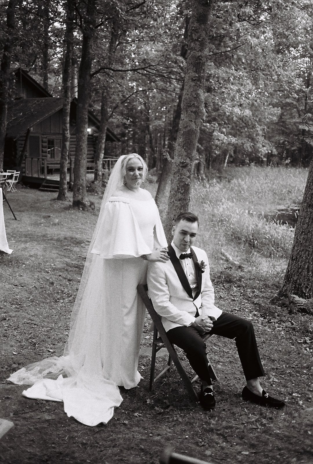 A bride in a long white wedding dress and veil standing next to a groom in a tuxedo, seated on a chair in a wooded outdoor setting.