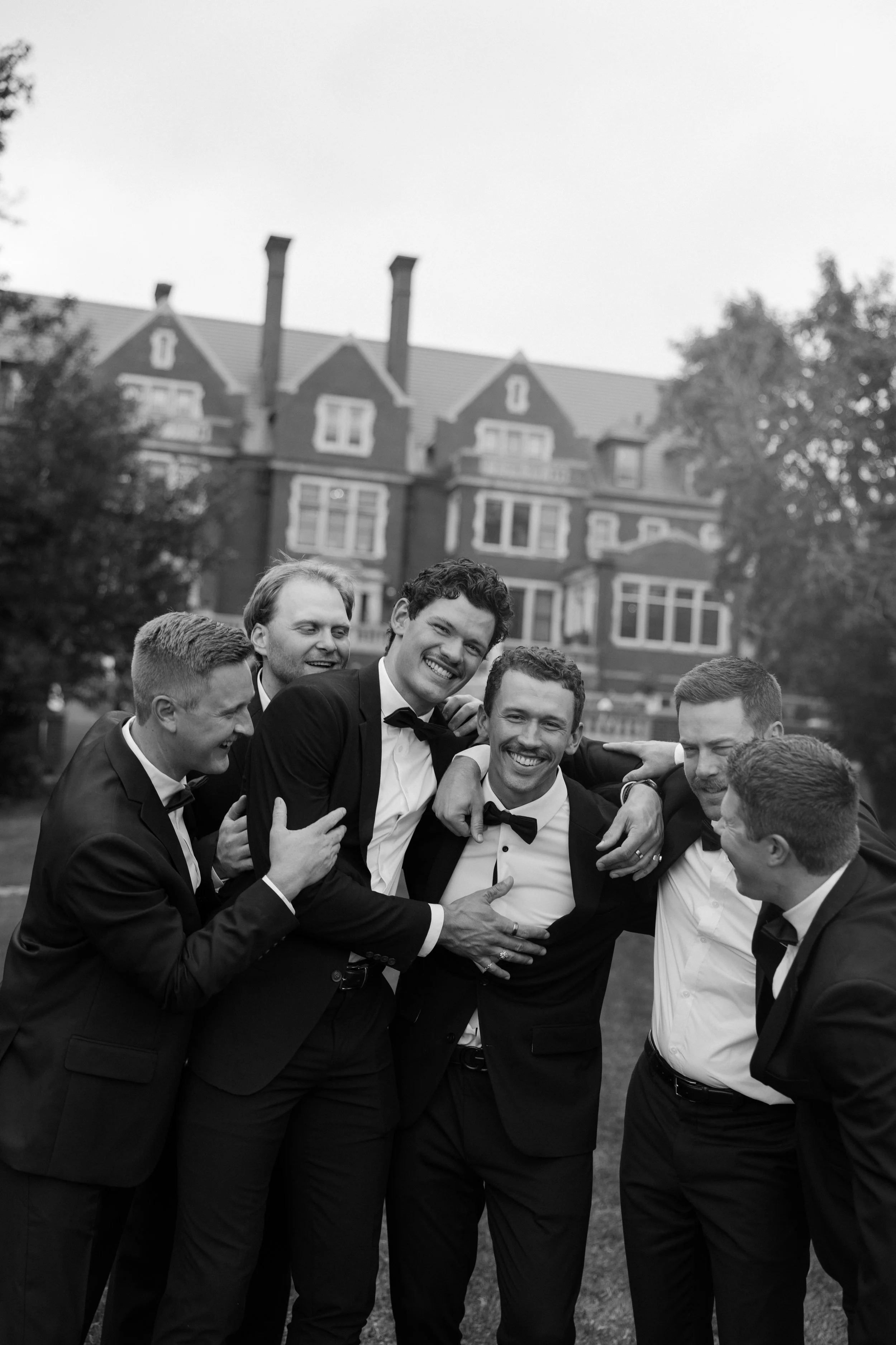 A group of men dressed in tuxedos and bow ties, smiling and gathering together outdoors in front of a large, historic brick house with multiple chimneys and trees in the background.