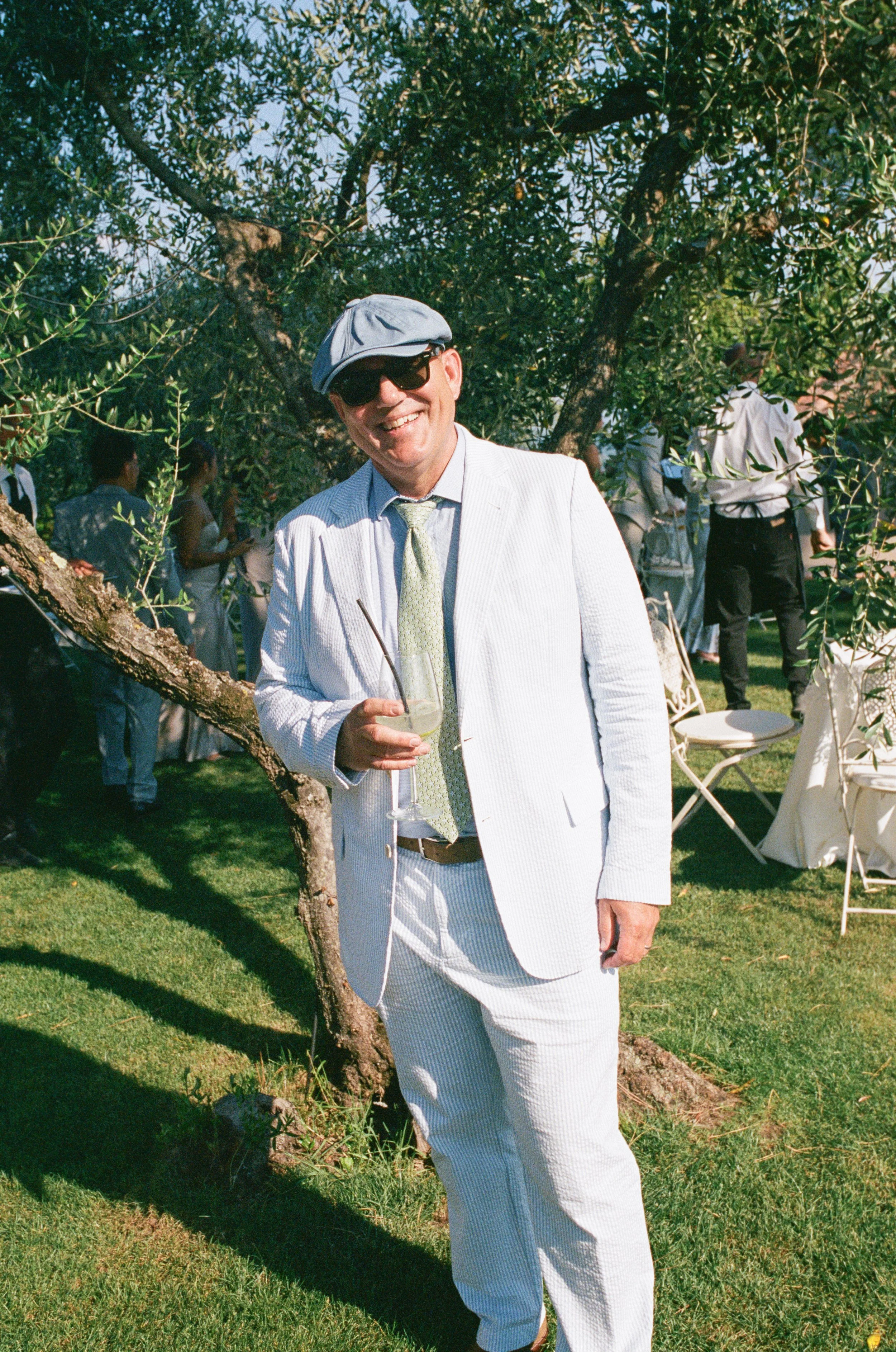 Man wearing a light-colored suit, striped shirt, and tie, holding a drink, standing under a tree at an outdoor gathering with other people in the background.