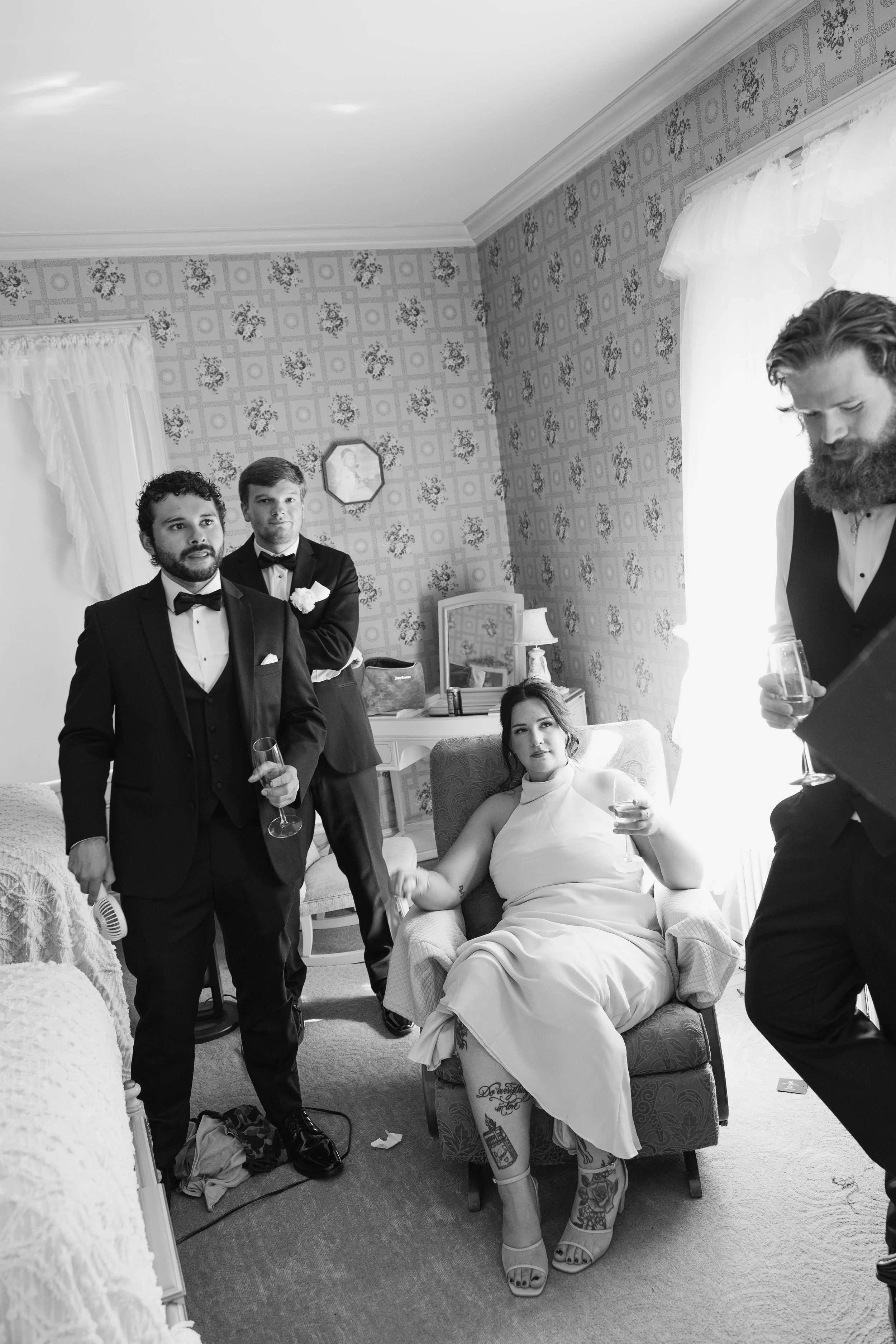 Group of well-dressed people at a formal gathering in a vintage-style room, with men in tuxedos and a woman sitting in an armchair holding a glass of wine.