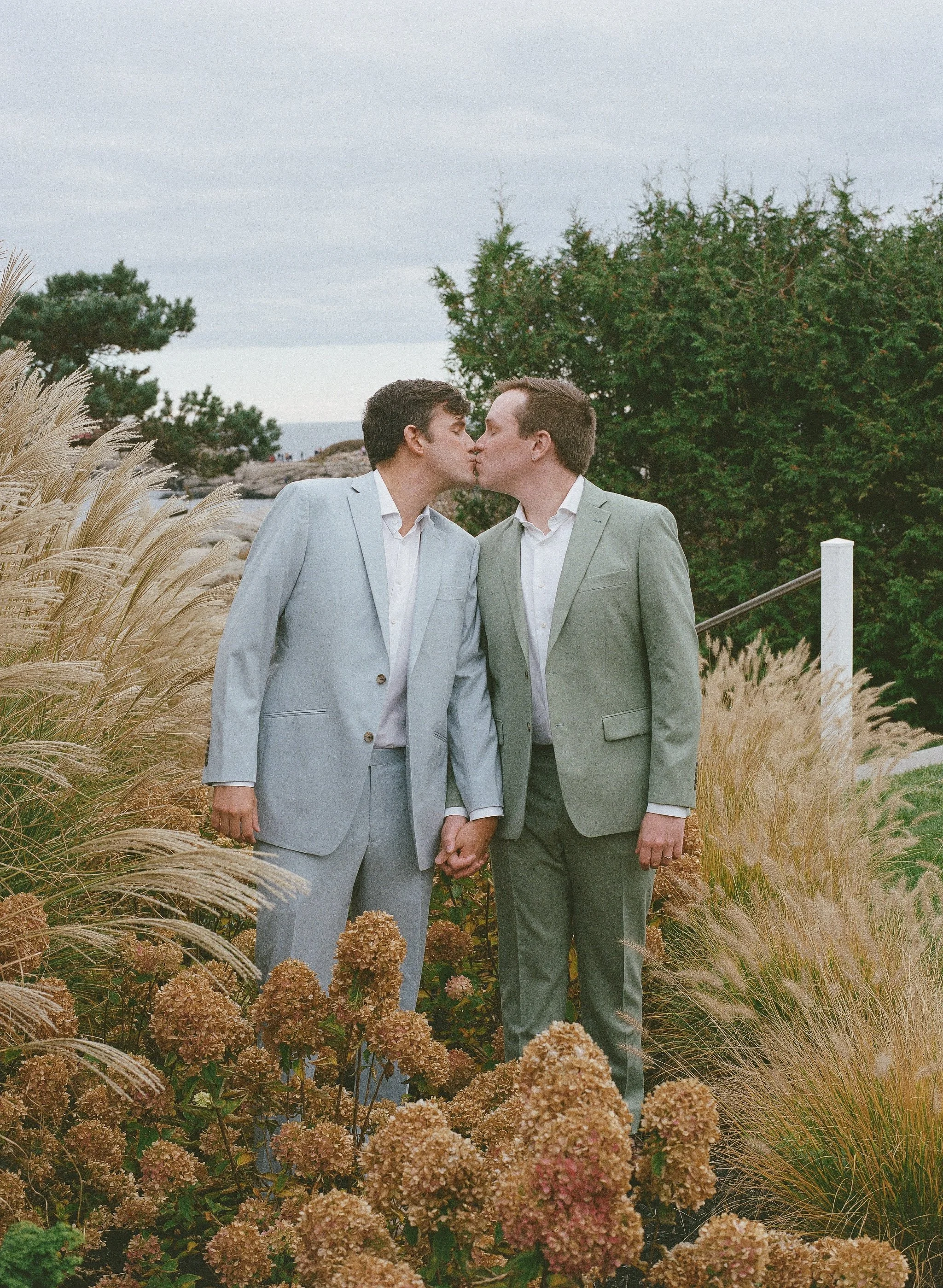 Two men dressed in suits kiss each other outdoors among tall grasses and flowers, with trees and cloudy sky in the background.