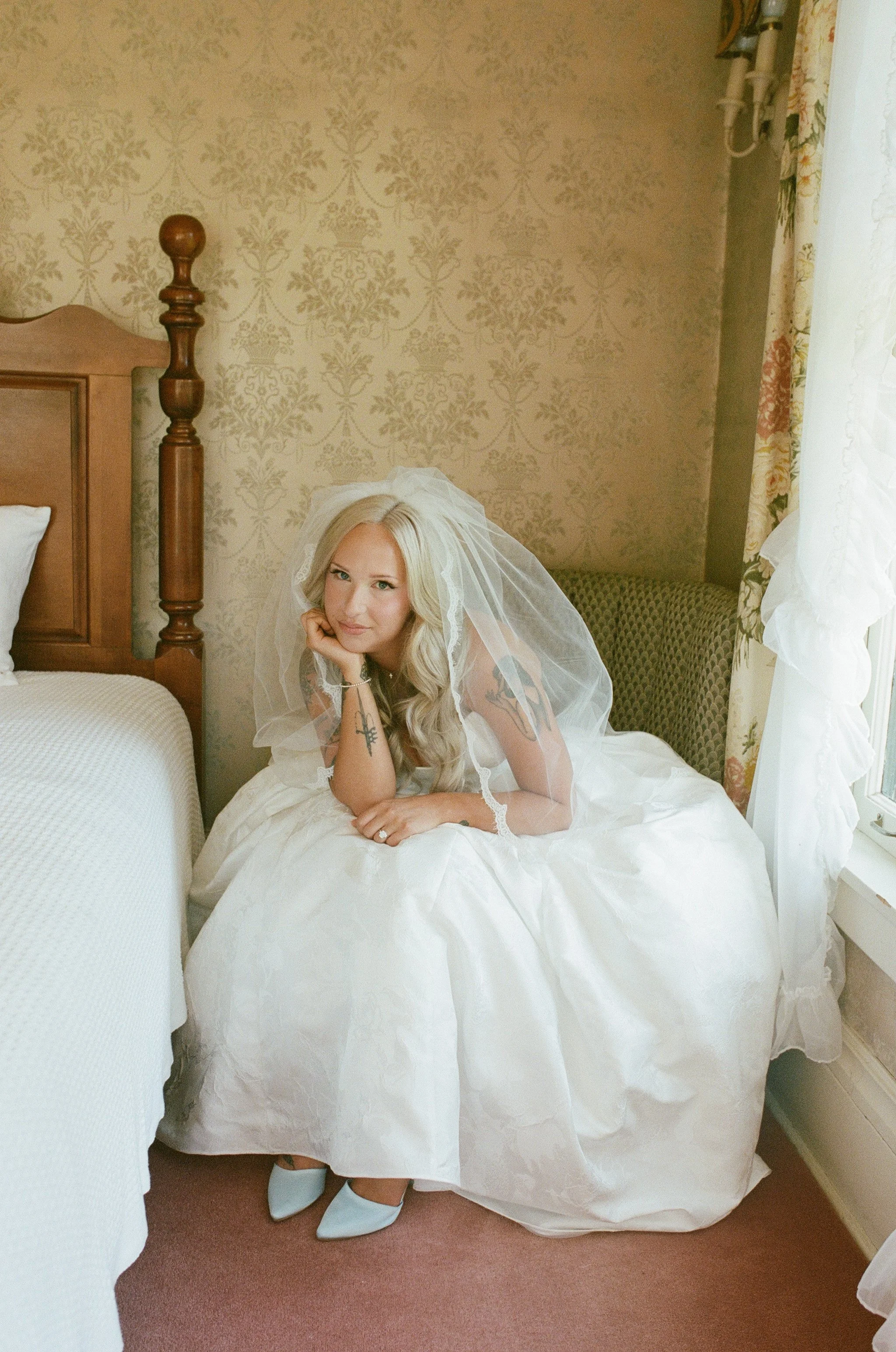 A woman with long blonde hair dressed in a wedding gown and veil, lying on a bed with her head resting on her hand, in a warmly decorated bedroom.
