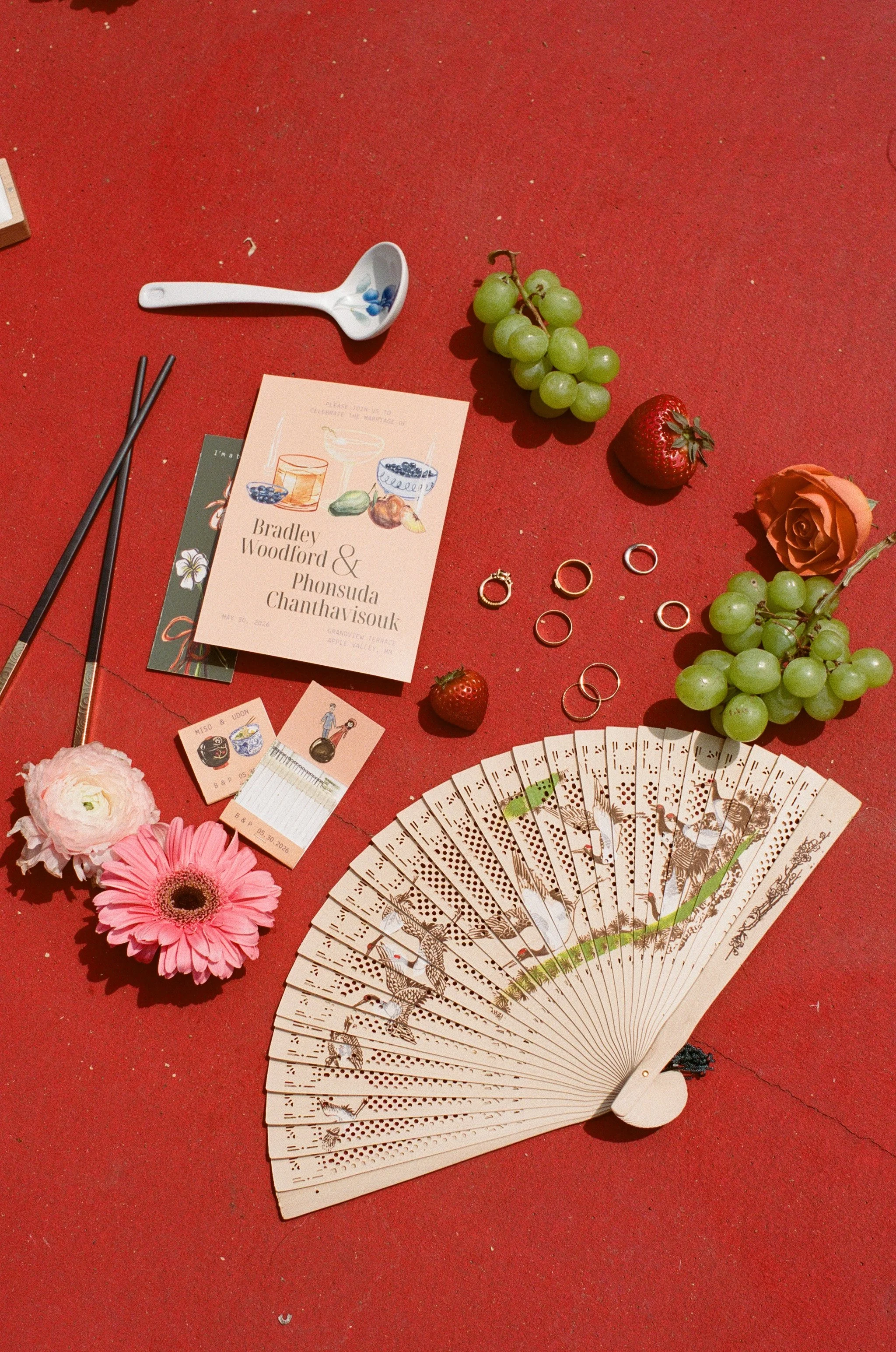 A red surface with various items including a white spoon with blue beans inside, two bubbles of grapes, a strawberry, a peach-colored rose, a pink flower, a fan with a scenic design, wedding rings, a wedding invitation, a small notebook, paper cards,