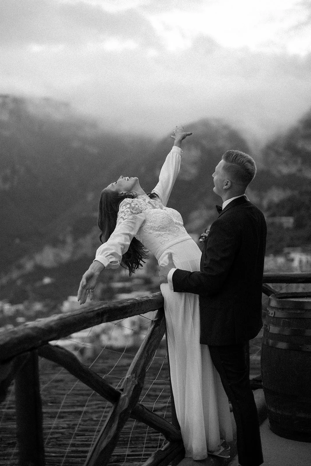 A black and white photo of a couple in wedding attire on a balcony with mountains in the background. The woman is leaning backward with her arm extended, while the man stands beside her holding her waist.