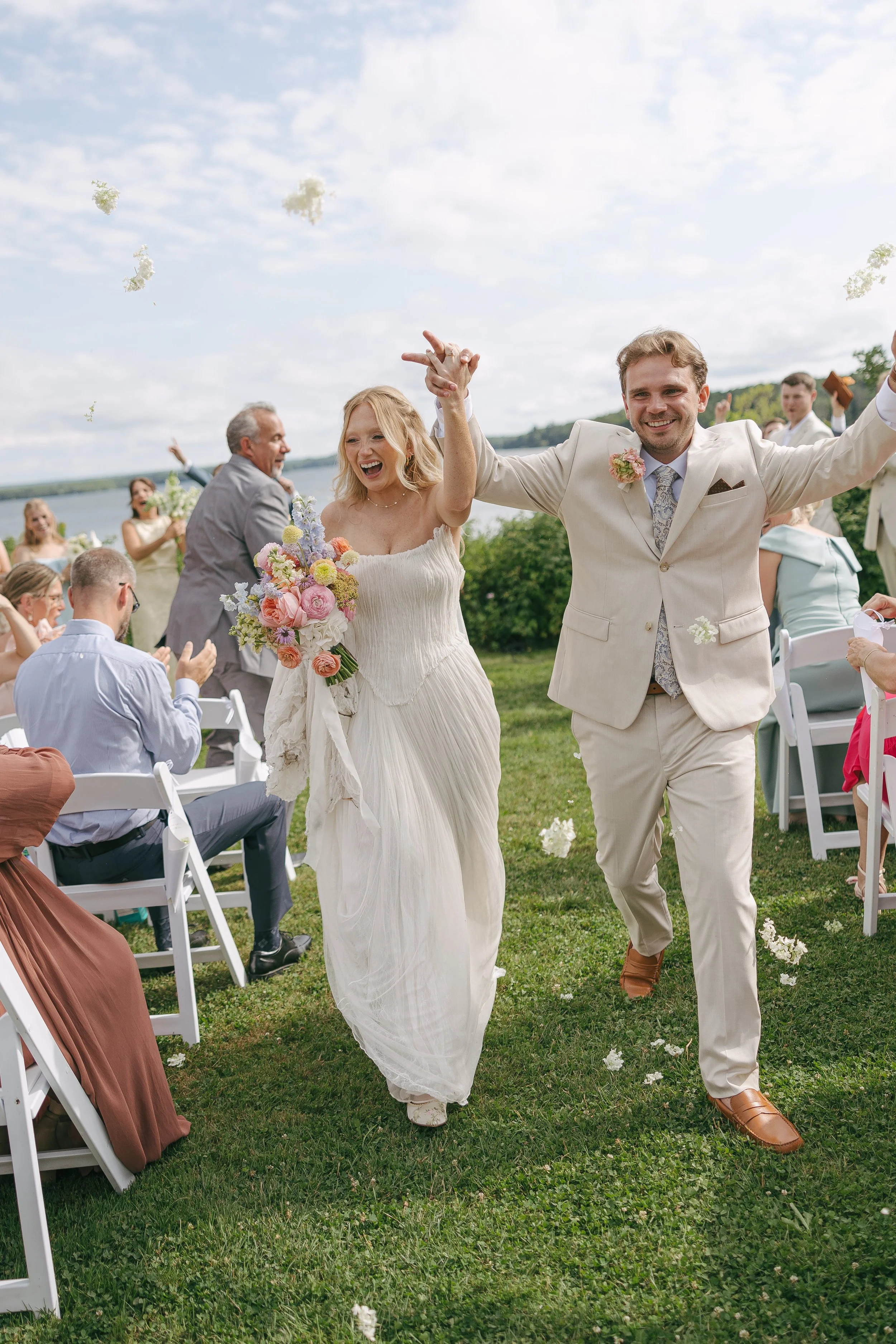 A newly married couple walking hand in hand outdoors, smiling and celebrating their wedding surrounded by guests, with a lake and trees in the background.