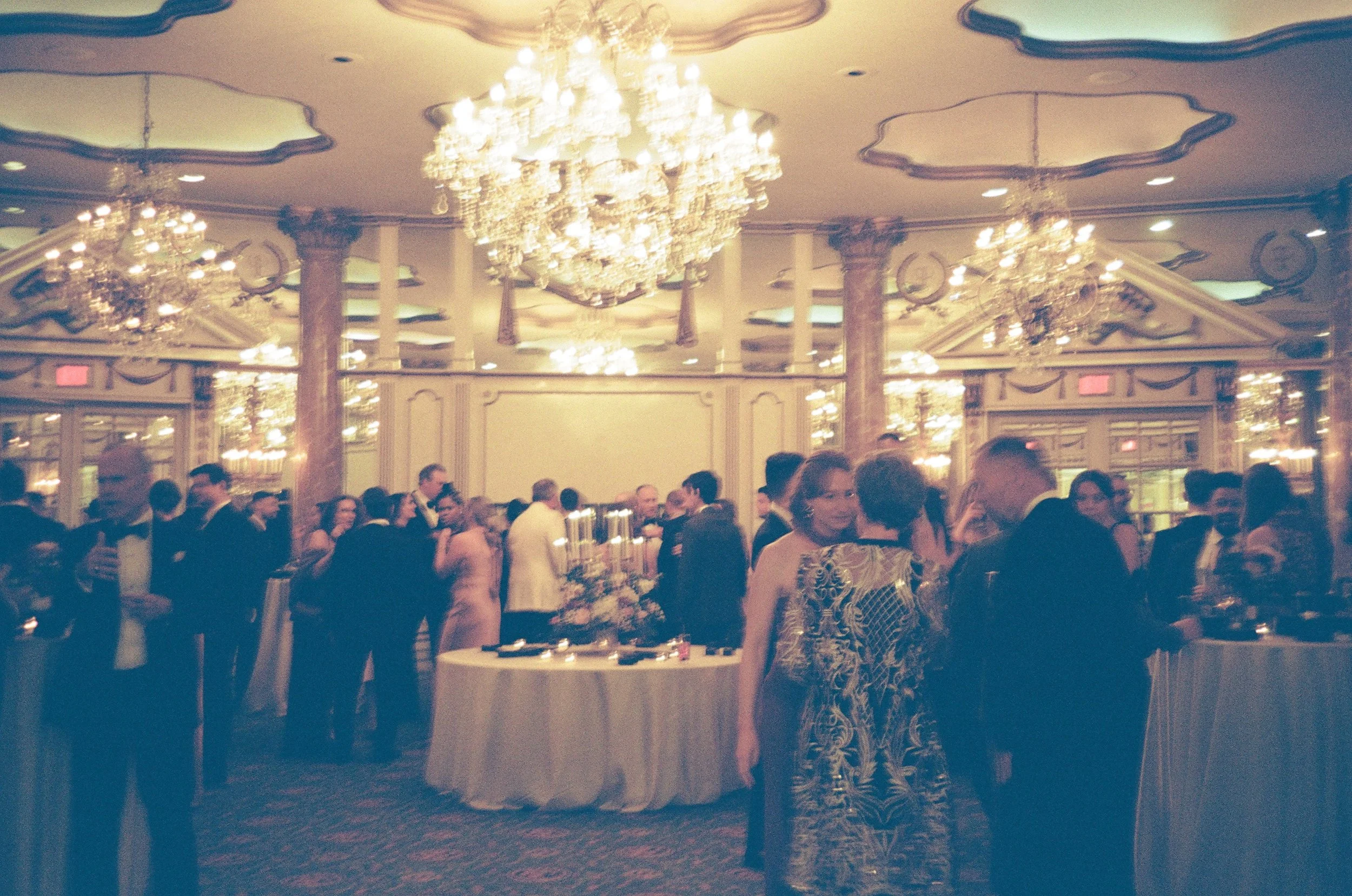 An elegant banquet hall with large crystal chandeliers hanging from an ornate ceiling, filled with well-dressed guests socializing at tables with white tablecloths.