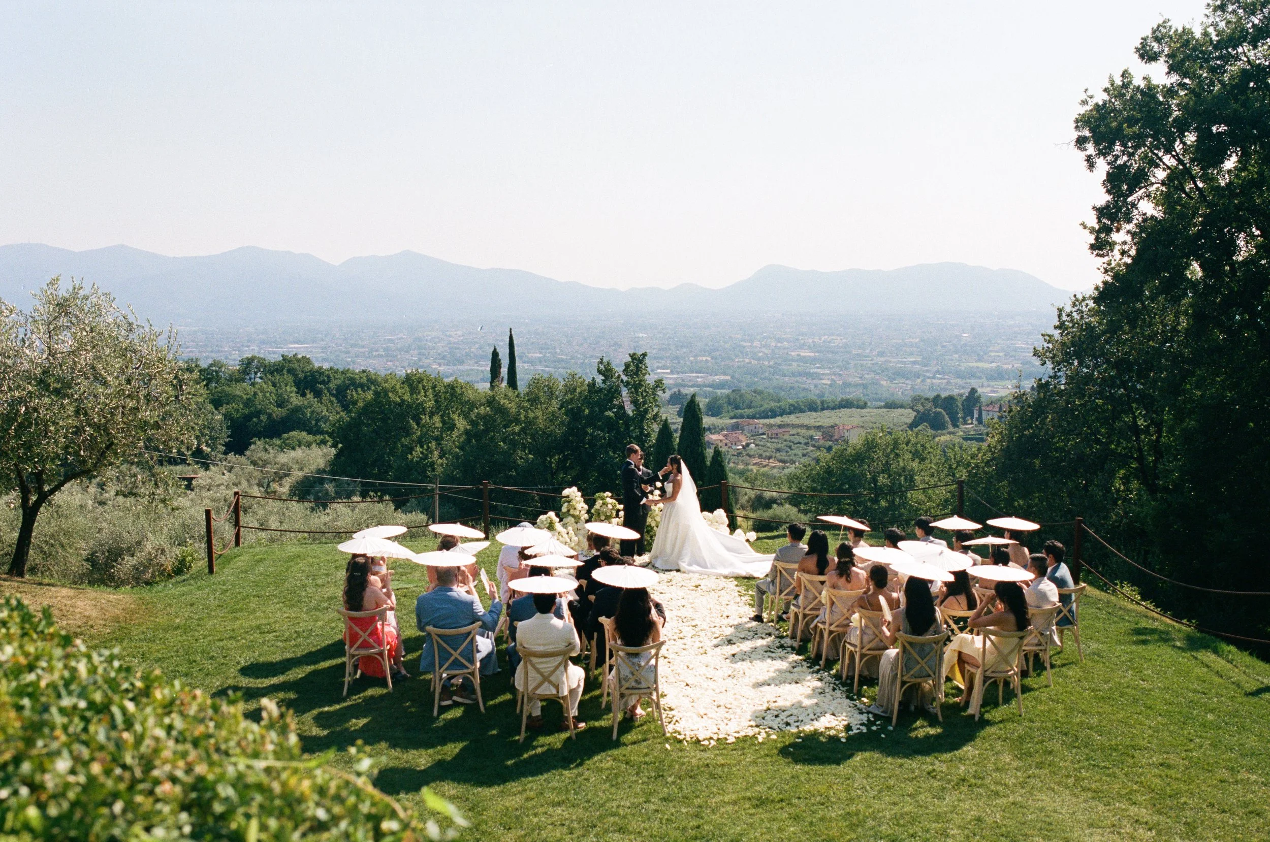Wedding ceremony outdoors with bride and groom exchanging vows, guests seated under umbrellas, scenic mountain view in the background.