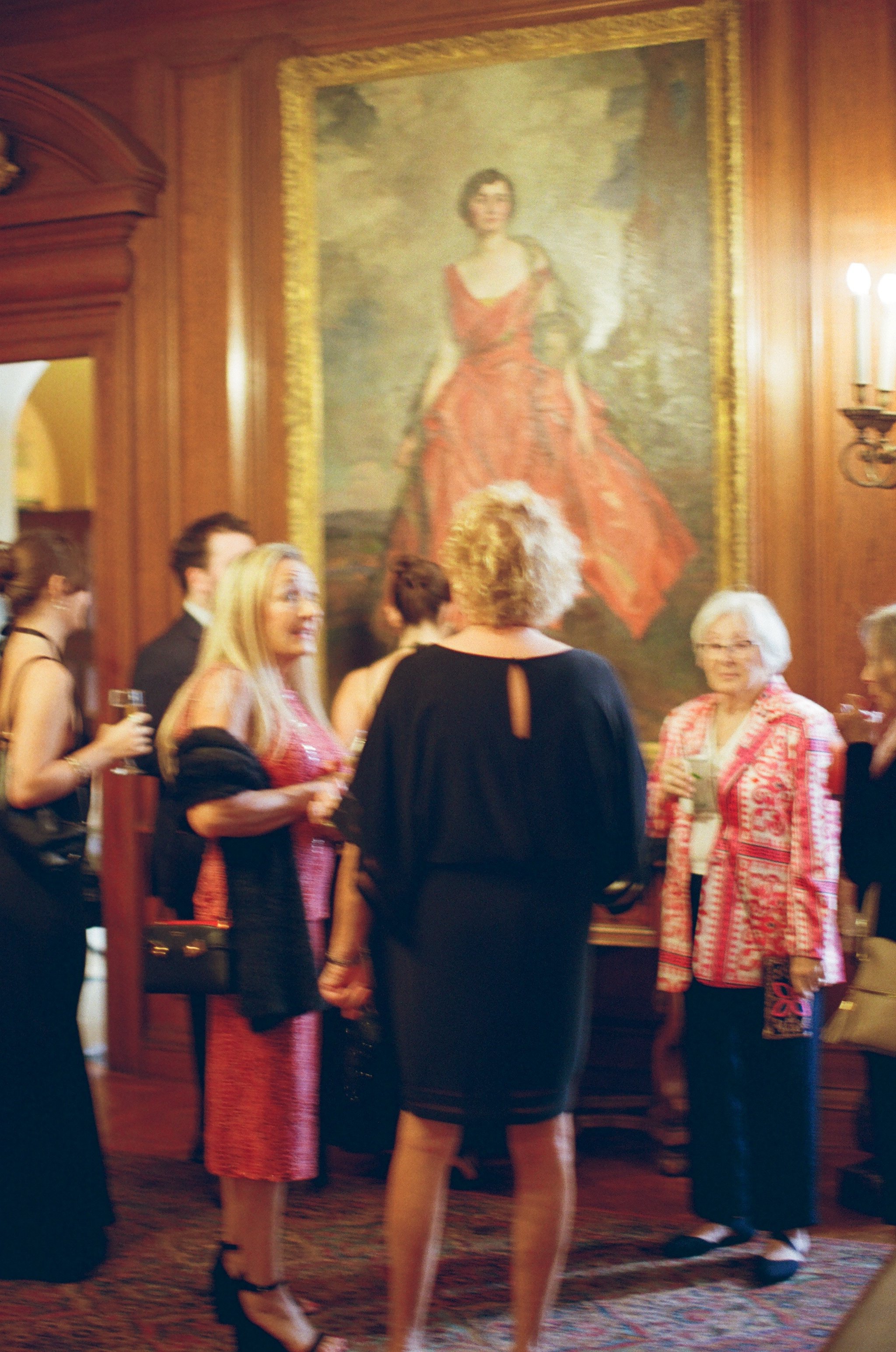A group of women dressed in formal and semi-formal attire standing in front of a large portrait painting of a woman in a red dress inside a wood-paneled room.