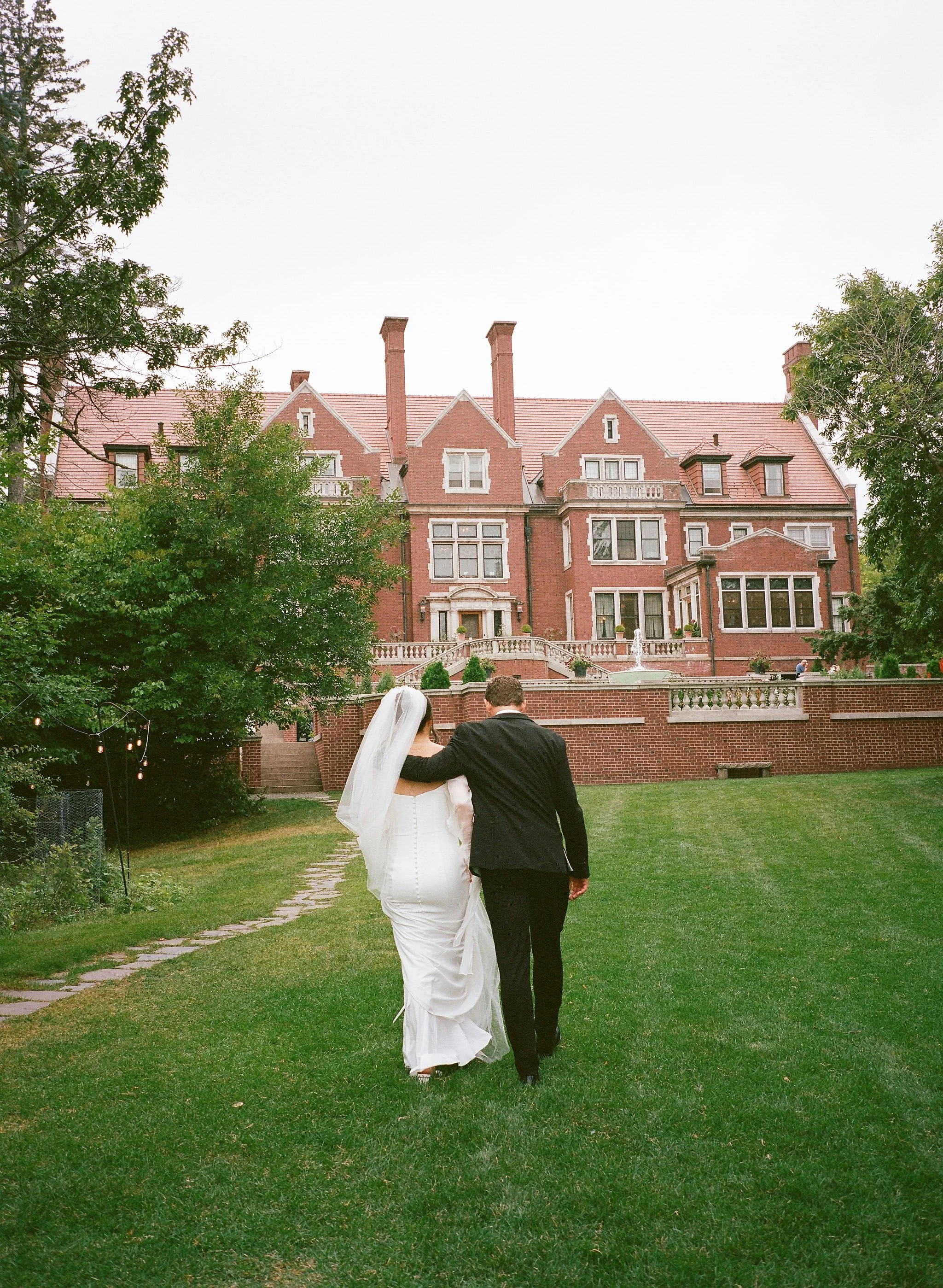 A bride and groom walking arm in arm on a grassy lawn with a large red brick mansion in the background.