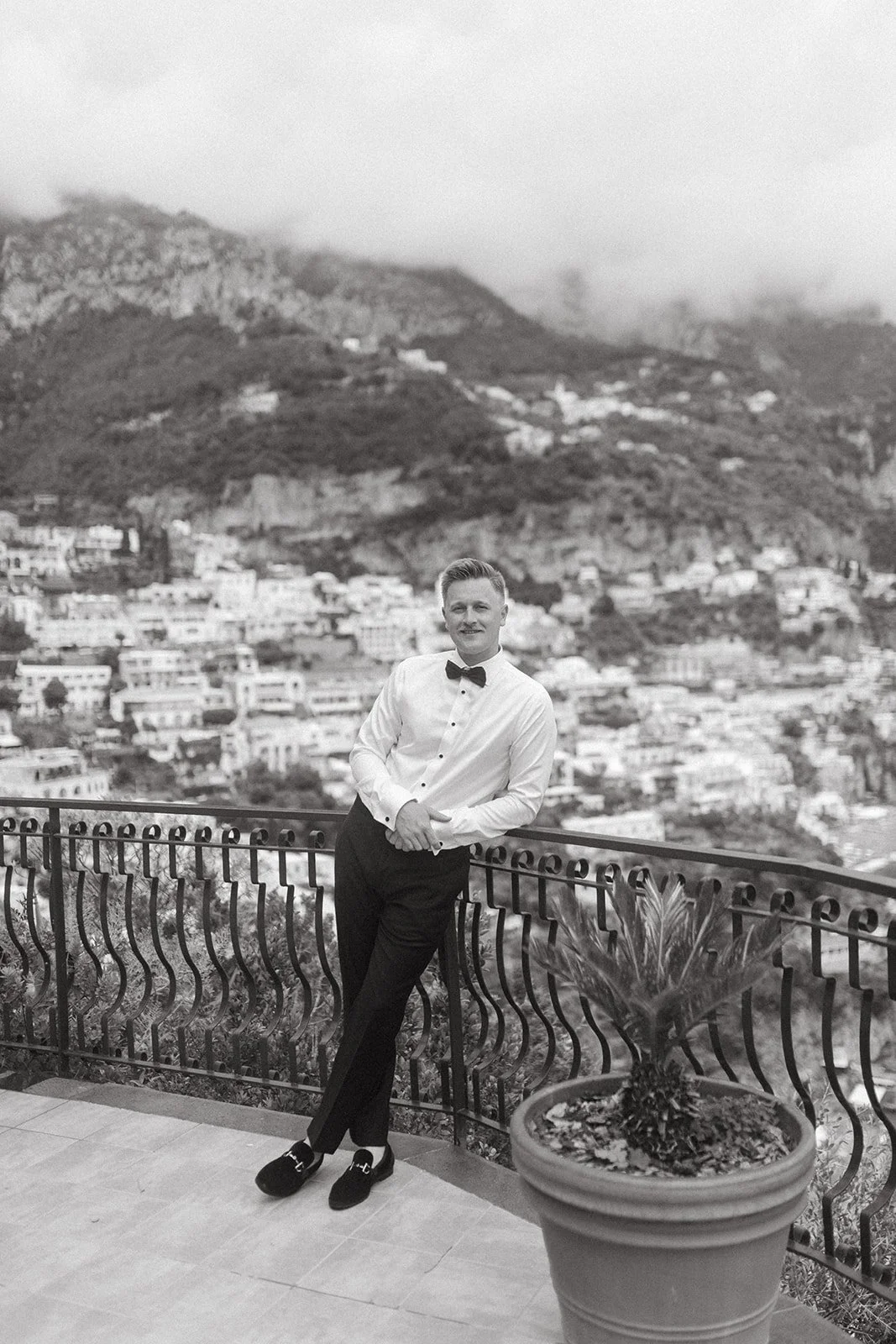 A man dressed in formal attire with a bow tie, standing on a balcony with a cityscape and mountains in the background.