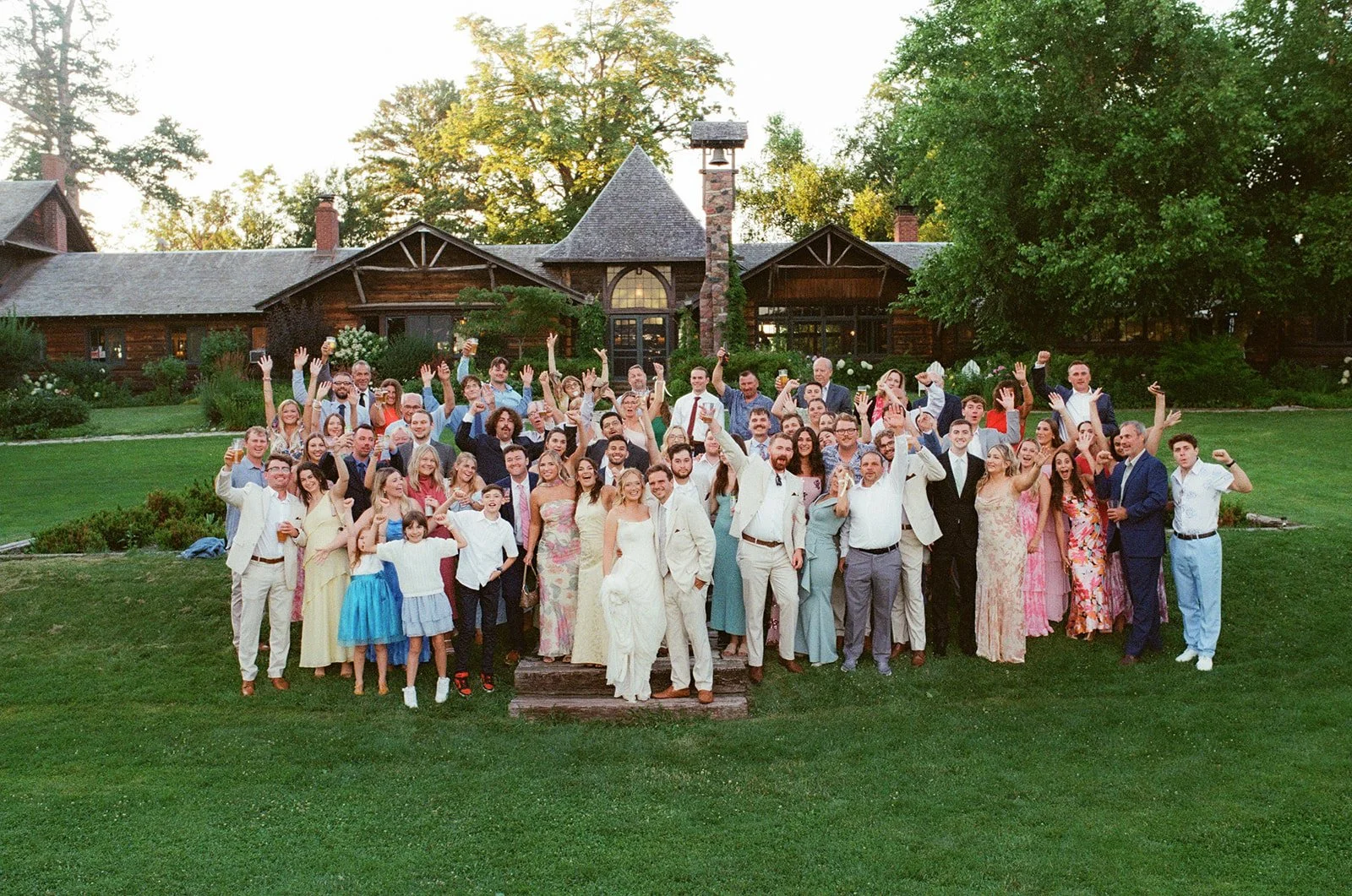 A large group of people gathered outdoors in front of a rustic wooden house, celebrating with many holding drinks and smiling, during a festive event.