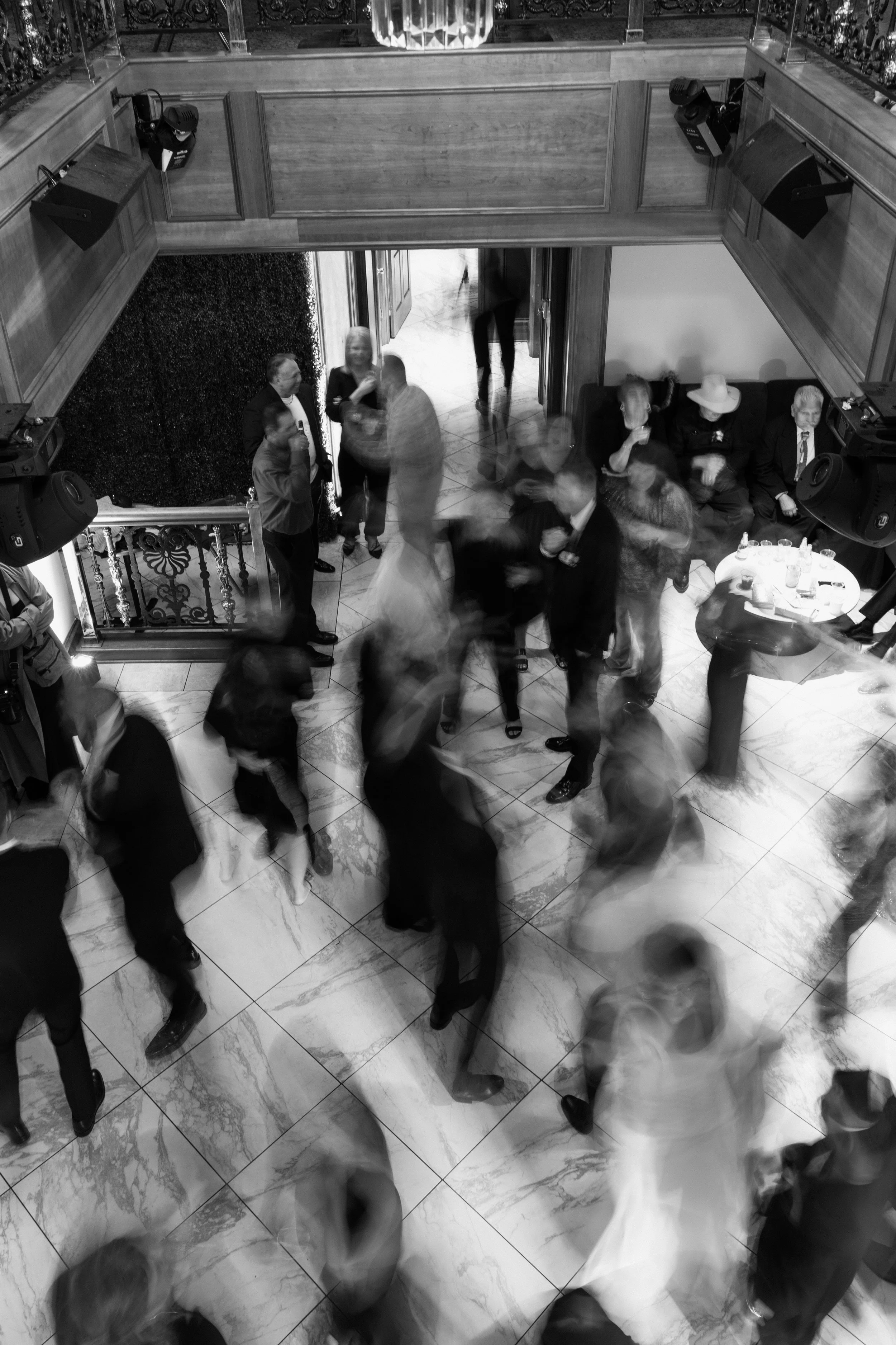 People socializing in a large, elegant indoor space with marble floors and wooden paneling on the walls, some standing and talking, others seated, with motion blur indicating movement.