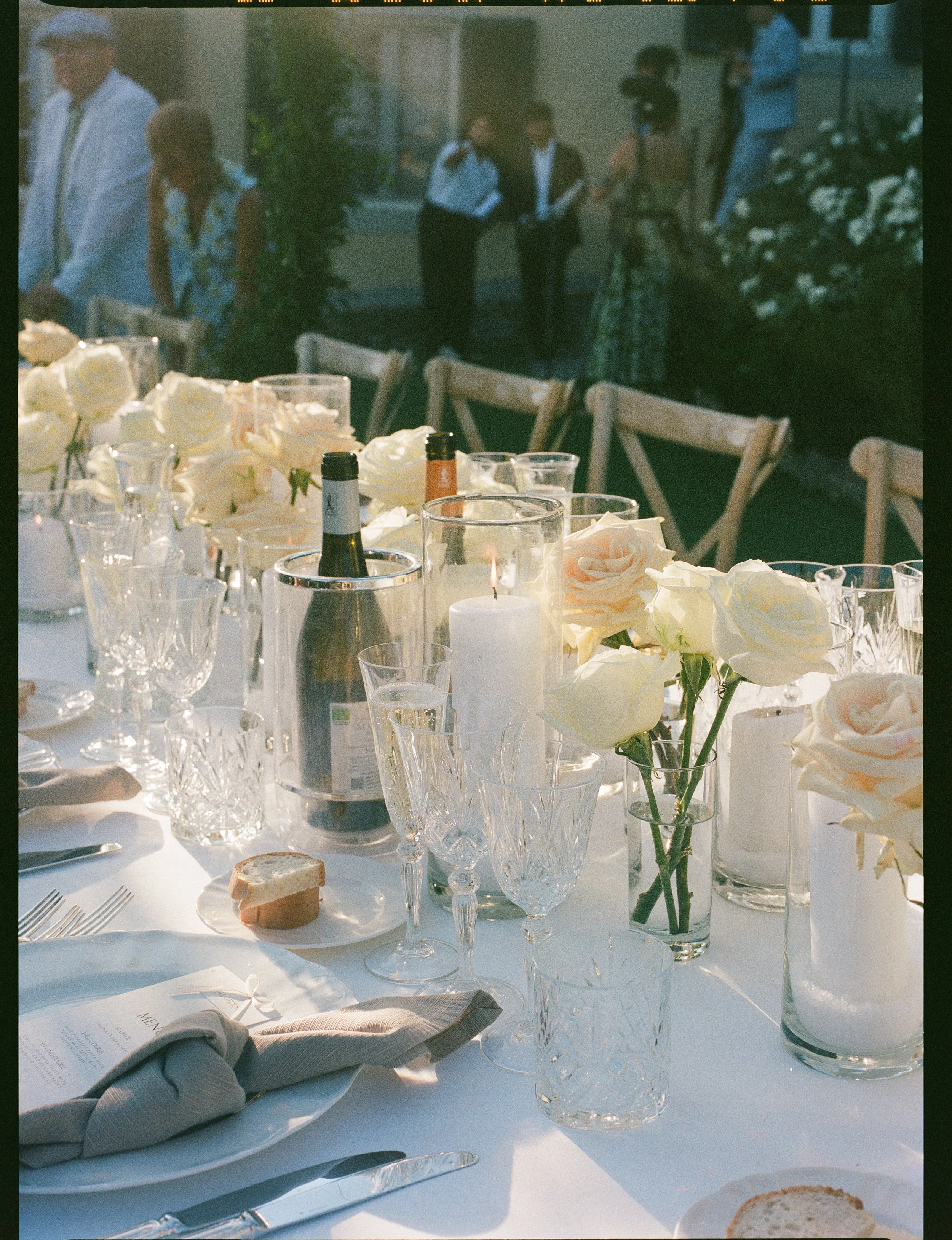 A formal outdoor table setting for a celebration with a white tablecloth, transparent glassware, white roses in vases, a white candle, bread slices, napkins, and silverware. In the background, there are people standing and talking, with some reading 