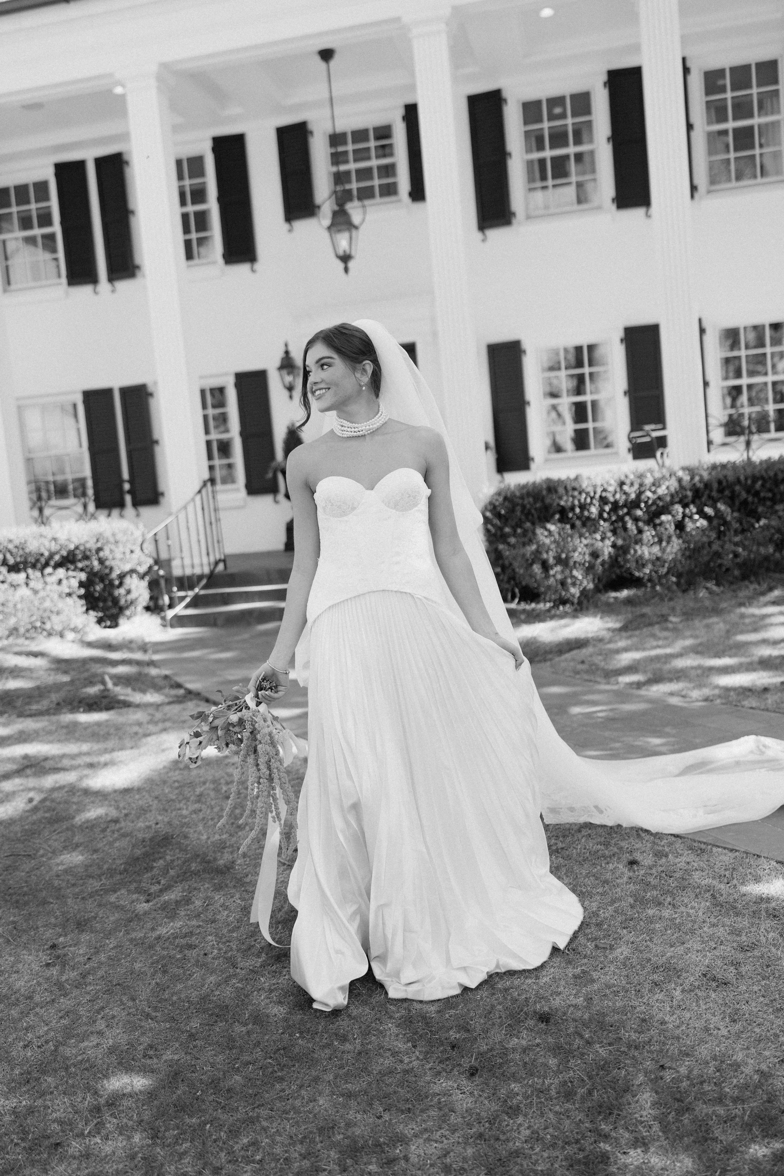 Black and white photo of a bride in a strapless wedding dress and pearl necklace, holding a bouquet, standing outdoors in front of a white house with dark shutters and windows, shrubbery, and stairs.