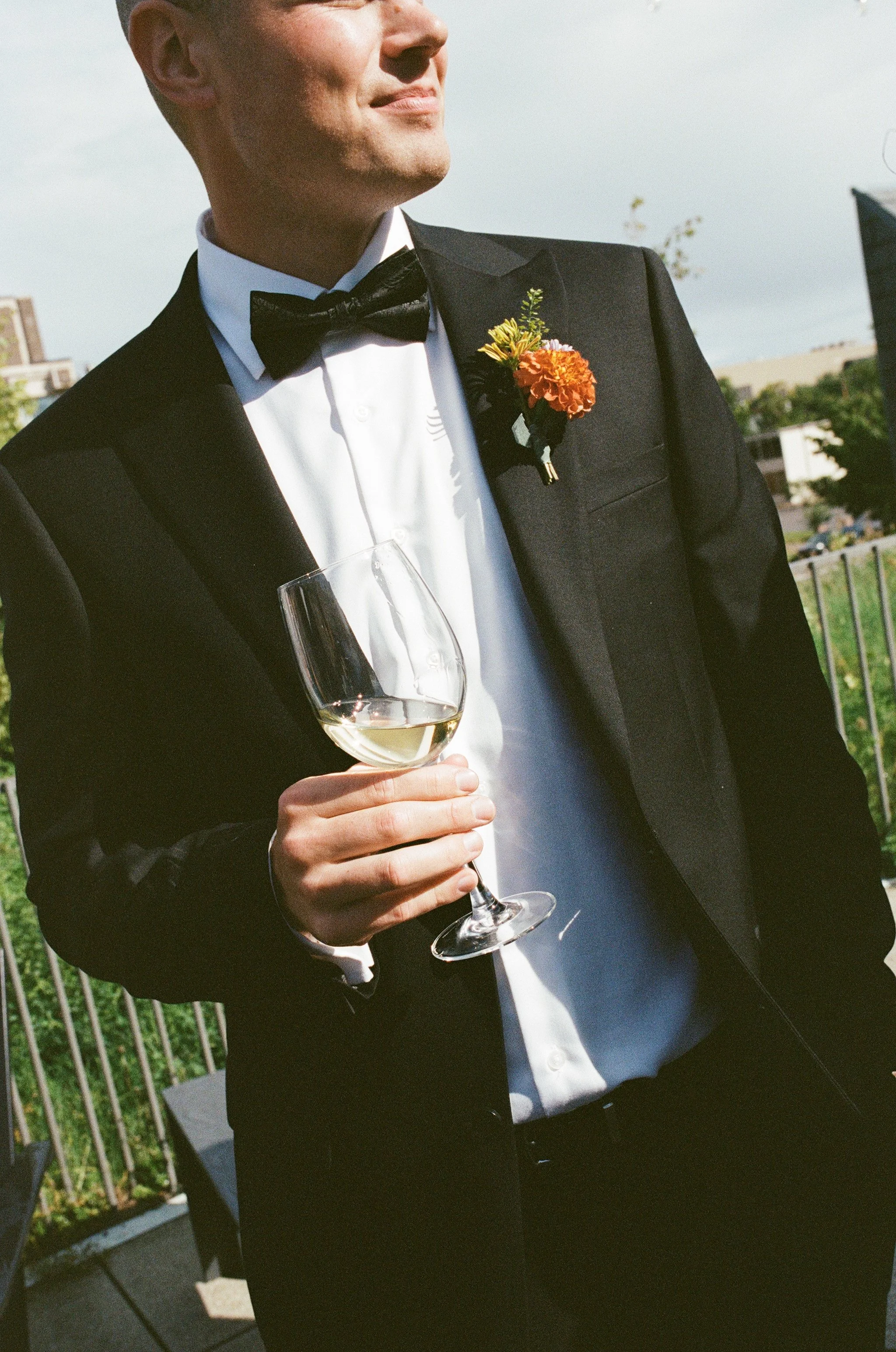 A man in a tuxedo with a boutonniere, holding a glass of white wine, standing outdoors.