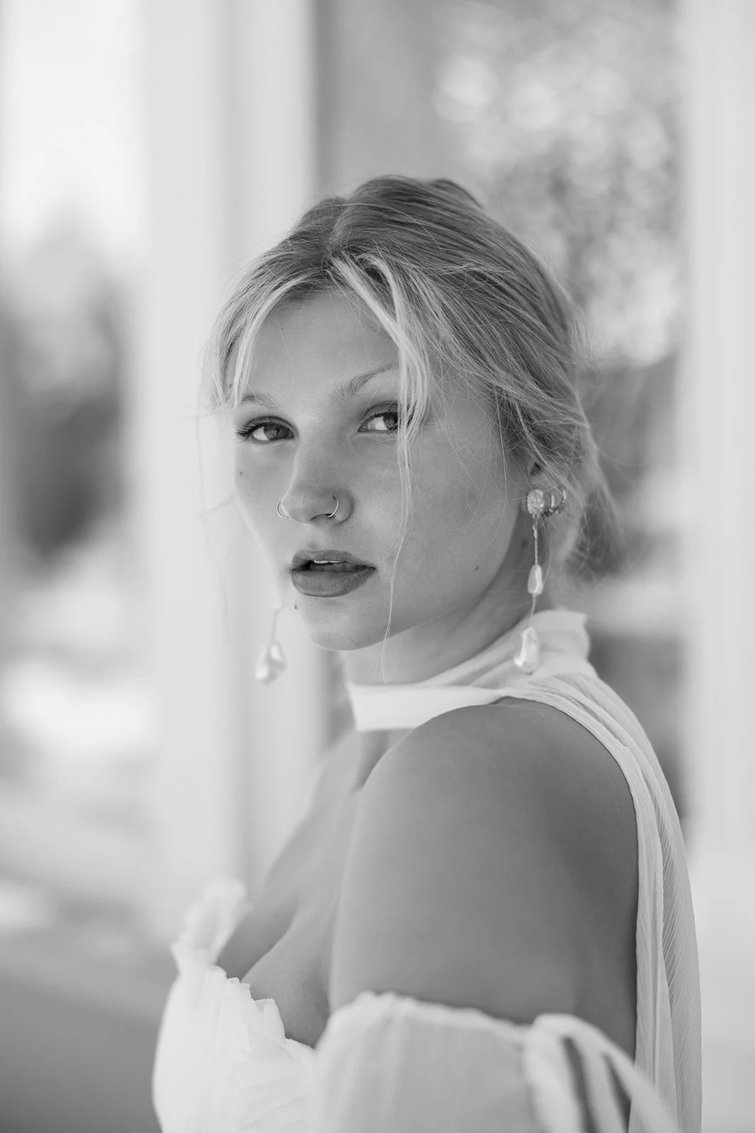 Black and white portrait of a young woman with blonde hair, wearing earrings, a nose ring, and a sleeveless top, looking at the camera with a slightly open mouth.
