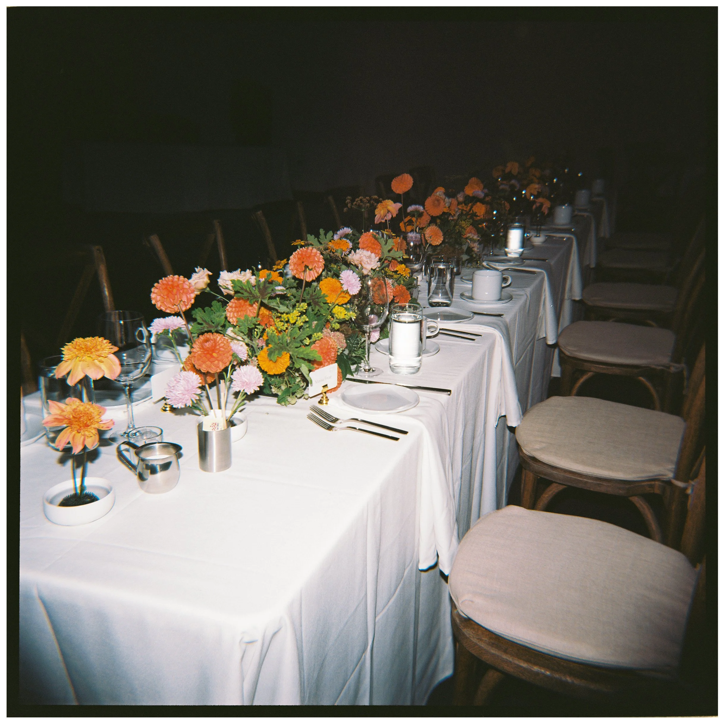 A long banquet table decorated with many colorful flower arrangements in vases and surrounded by chairs, set for a formal event or party.