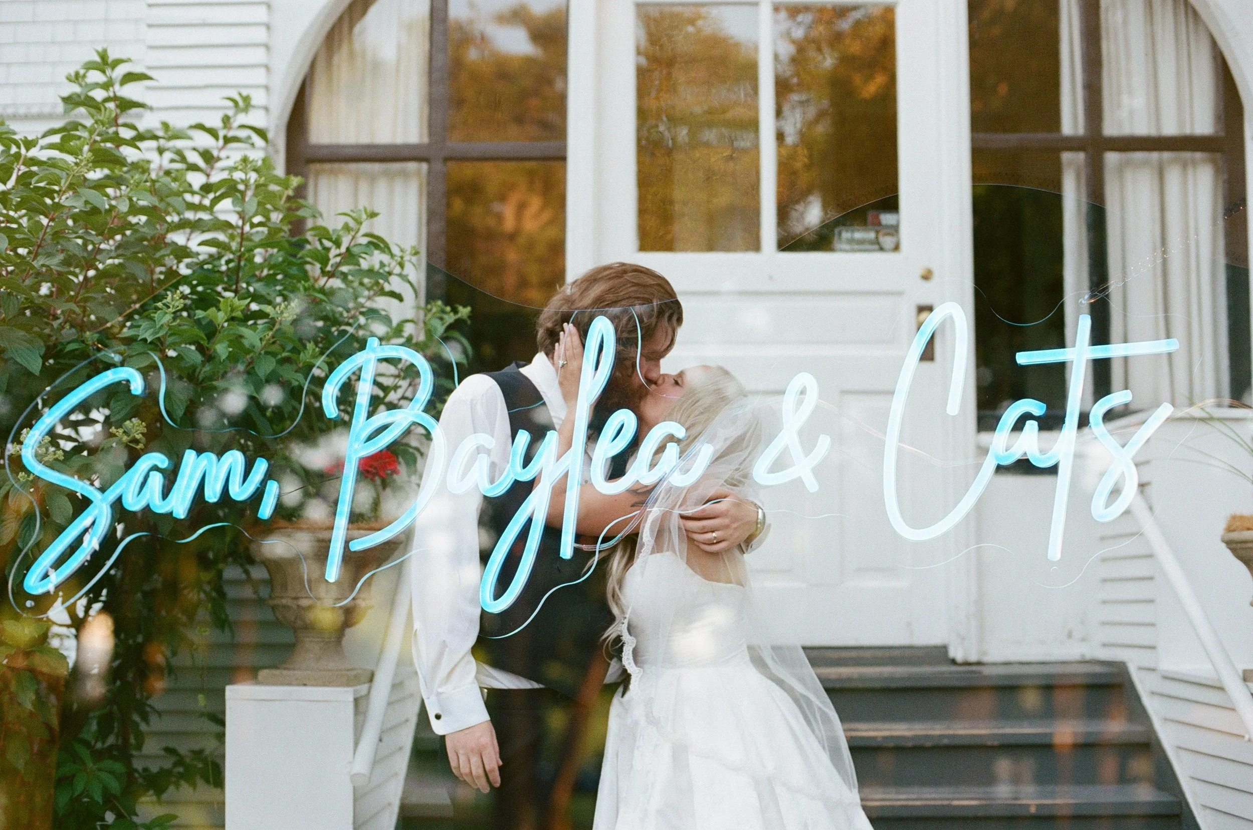 A couple kisses through a window with a neon sign that reads 'Sam, Paige & Clay' and some greenery outside.