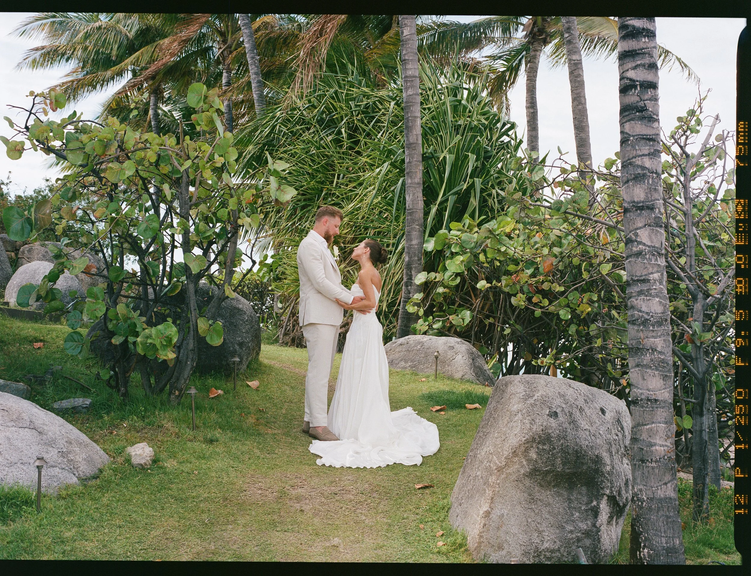 A couple dressed in white, the man in a suit and the woman in a wedding dress, holding hands and looking at each other in a lush tropical outdoor setting with palm trees and large rocks.