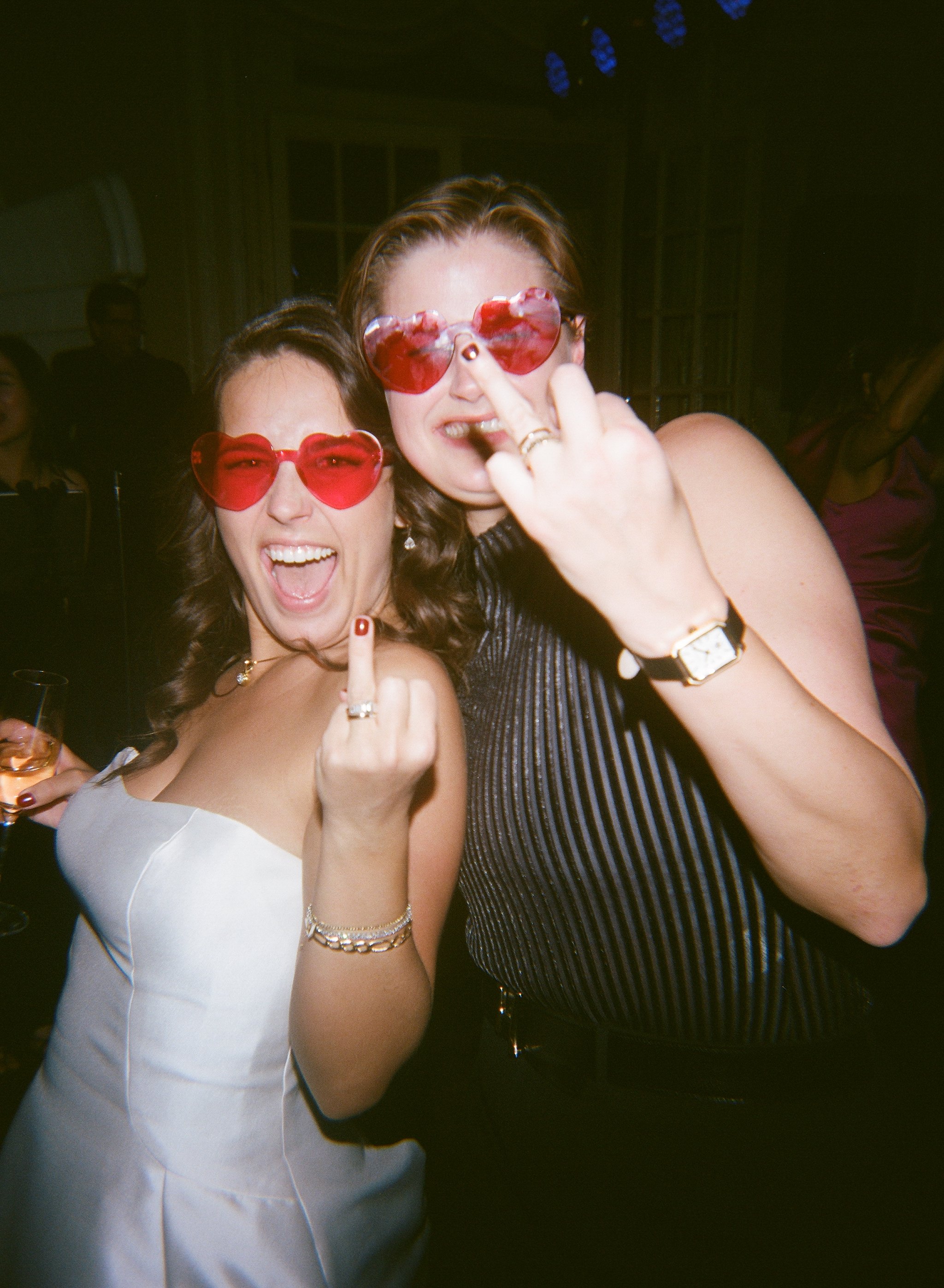 Two women in party attire wearing heart-shaped sunglasses smiling and making playful gestures at a celebration or party.