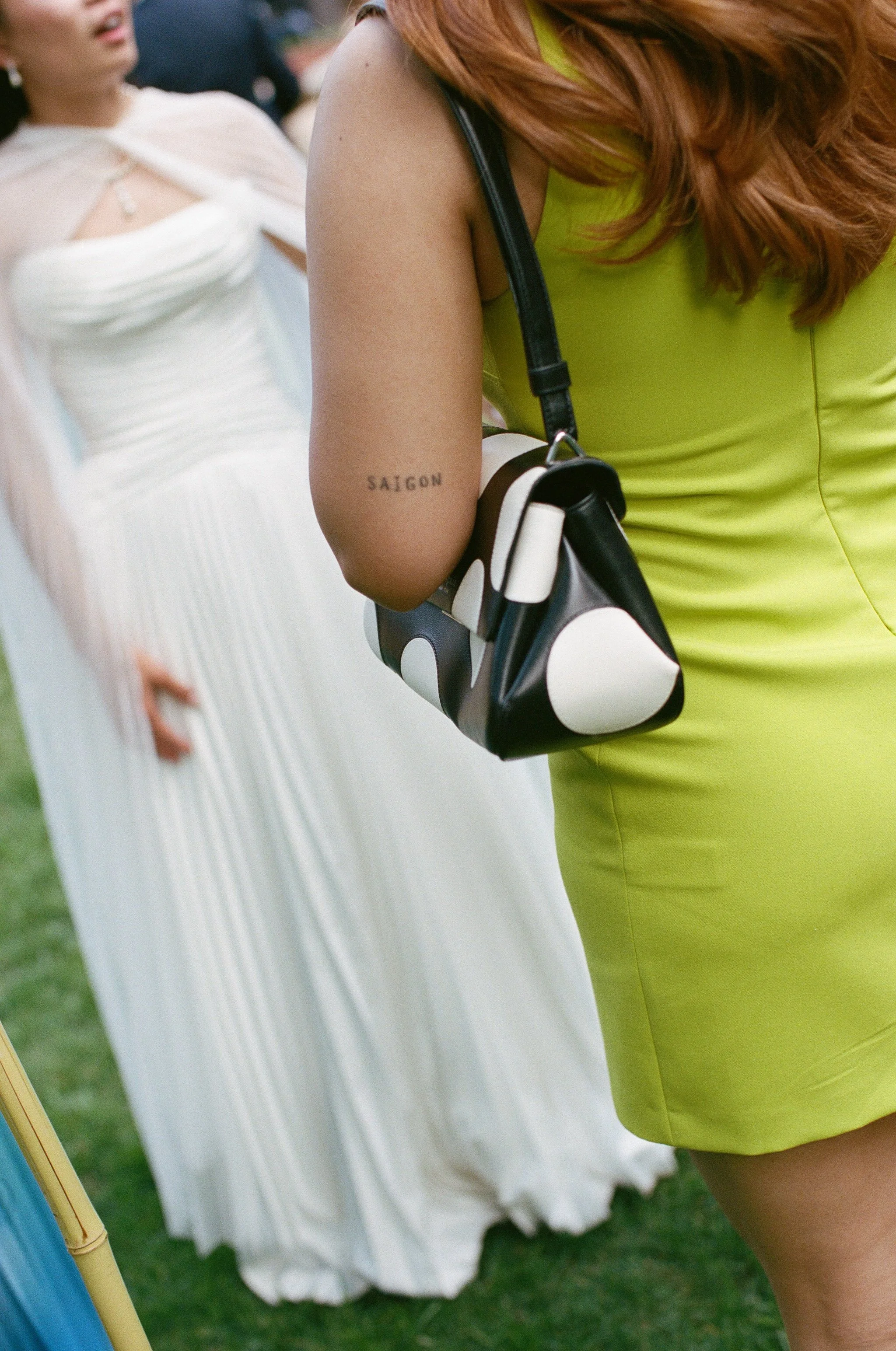 Close-up of a woman's arm with a tattoo reading 'SAIGON', wearing a lime green dress and holding a black-and-white polka dot handbag at an outdoor event. In the background, a person in a white dress is partially visible.