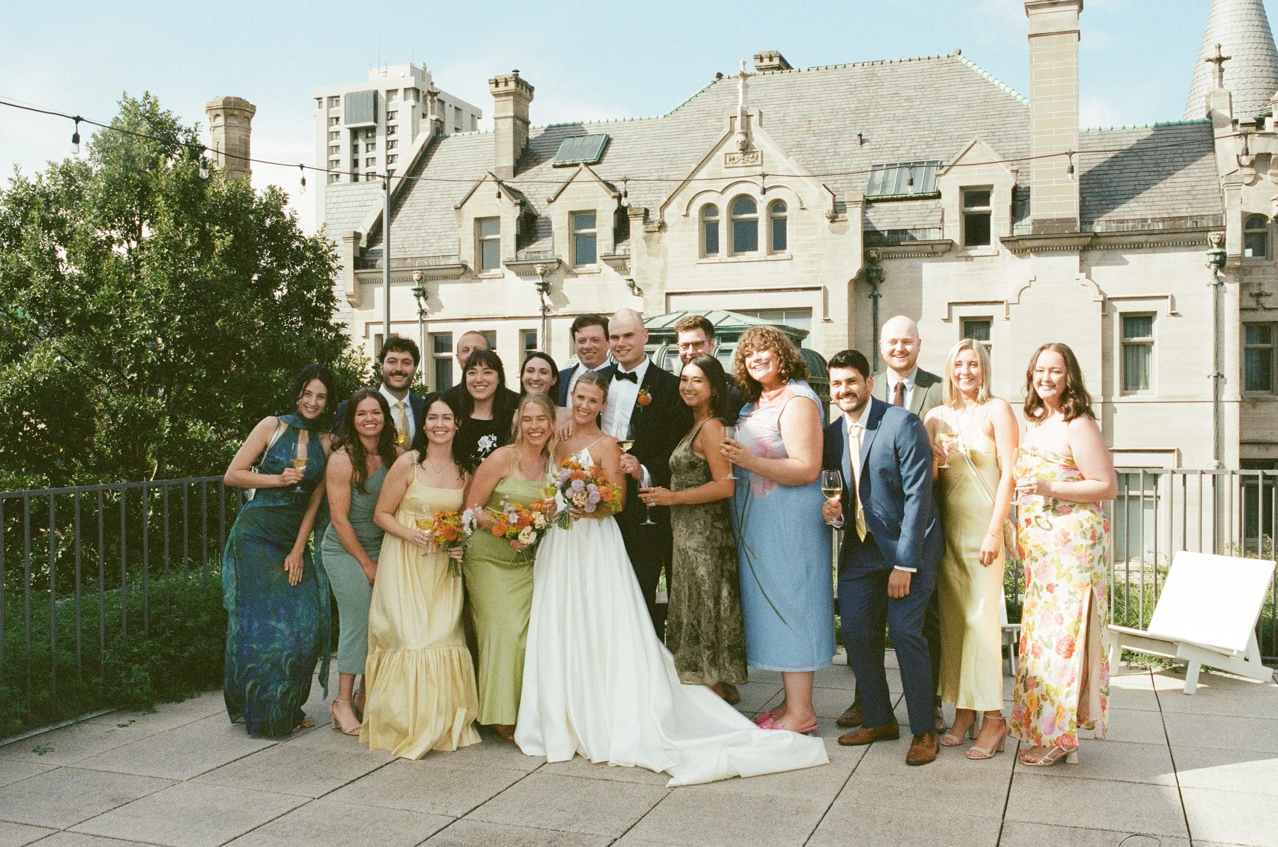 A group of people dressed in formal and colorful attire gathered outdoors in front of a large, historic mansion-like building for a celebration, likely a wedding, with some holding drinks and flowers.