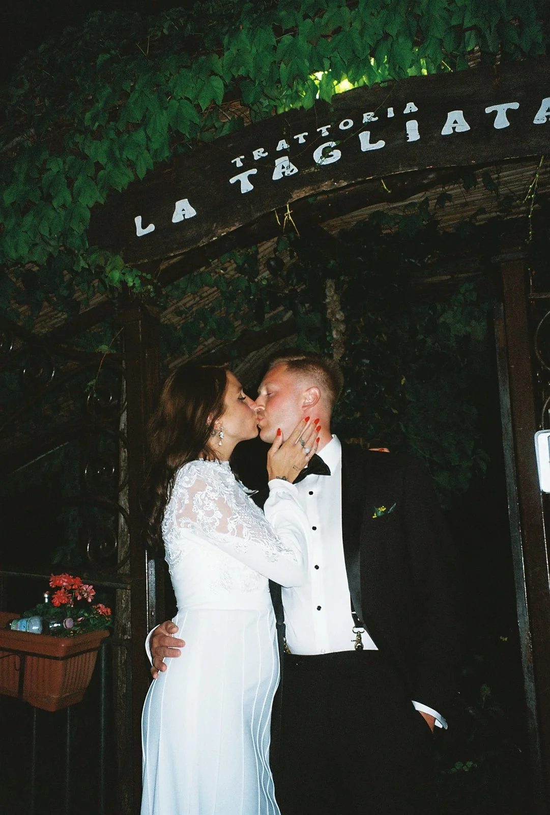 Bride and groom sharing a kiss at their wedding, standing under a wooden arch with green foliage. The bride is wearing a white lace dress, and the groom is in a black tuxedo with a bow tie.