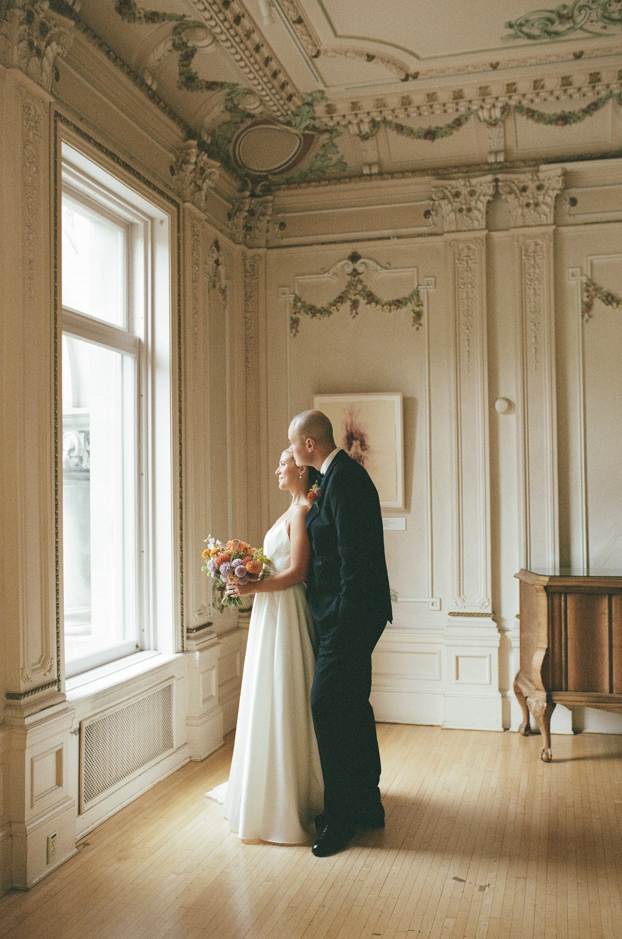 A bride and groom standing by a large window in a decorated room, with the bride holding a bouquet of flowers, and the groom leaning in close, both smiling gently.
