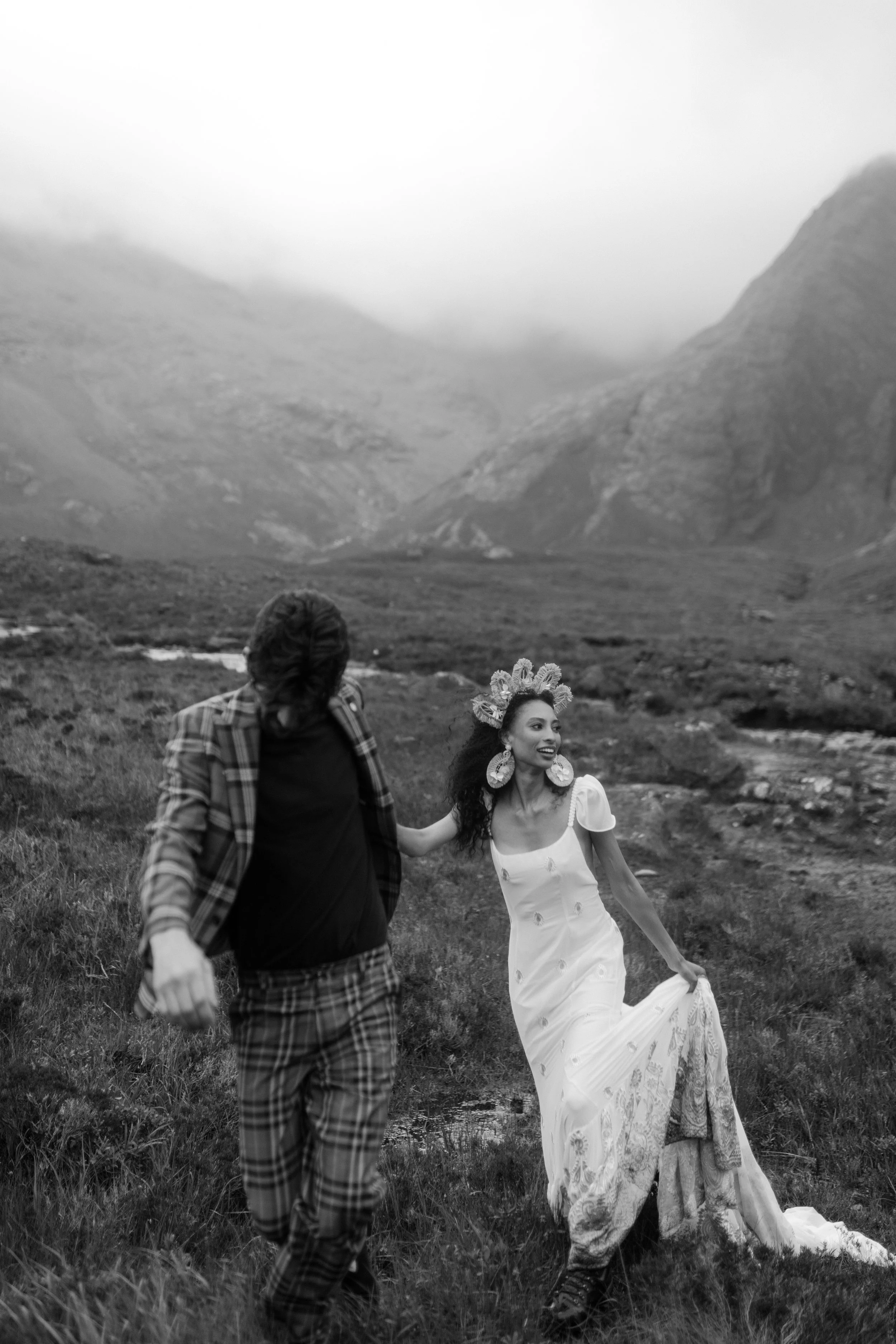 A man and a woman walking outdoors in a grassy field with mountains in the background. The woman is wearing a white dress with floral embroidery and large earrings, and the man is dressed in a plaid jacket and pants. The woman has a flower crown on her head.