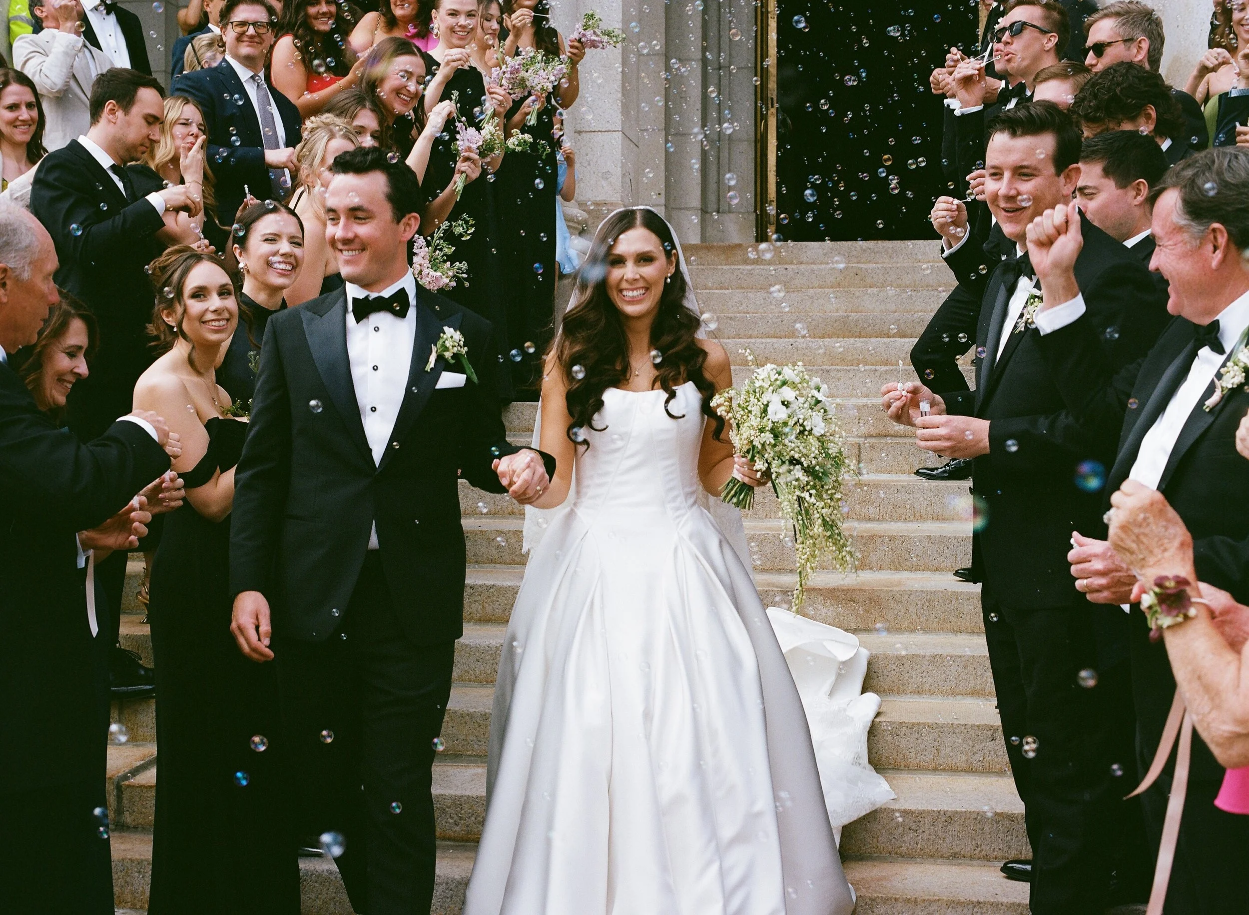 A woman in a white wedding dress holding a bouquet of flowers, smiling, walking down stairs at a wedding reception. She is surrounded by guests dressed in formal attire, blowing bubbles, and celebrating outdoors.