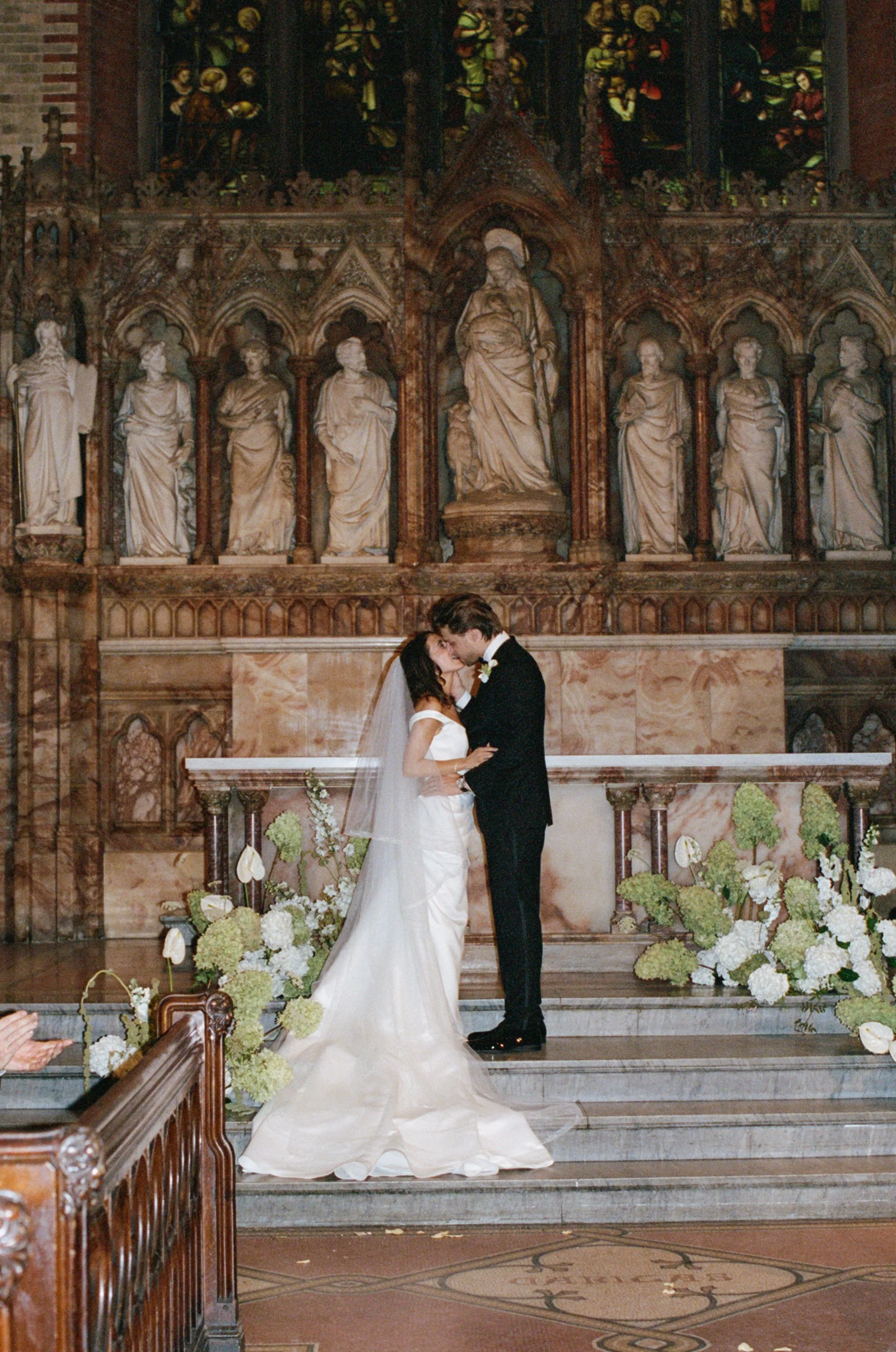 A bride and groom standing and kissing at the altar inside a church, with religious statues, stained glass windows, and floral arrangements surrounding them.