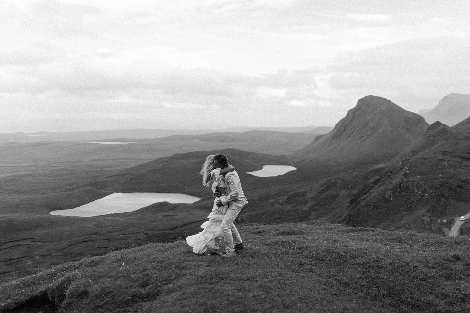 A black and white photo of a couple embracing in a scenic mountain landscape with lakes and hills in the background.